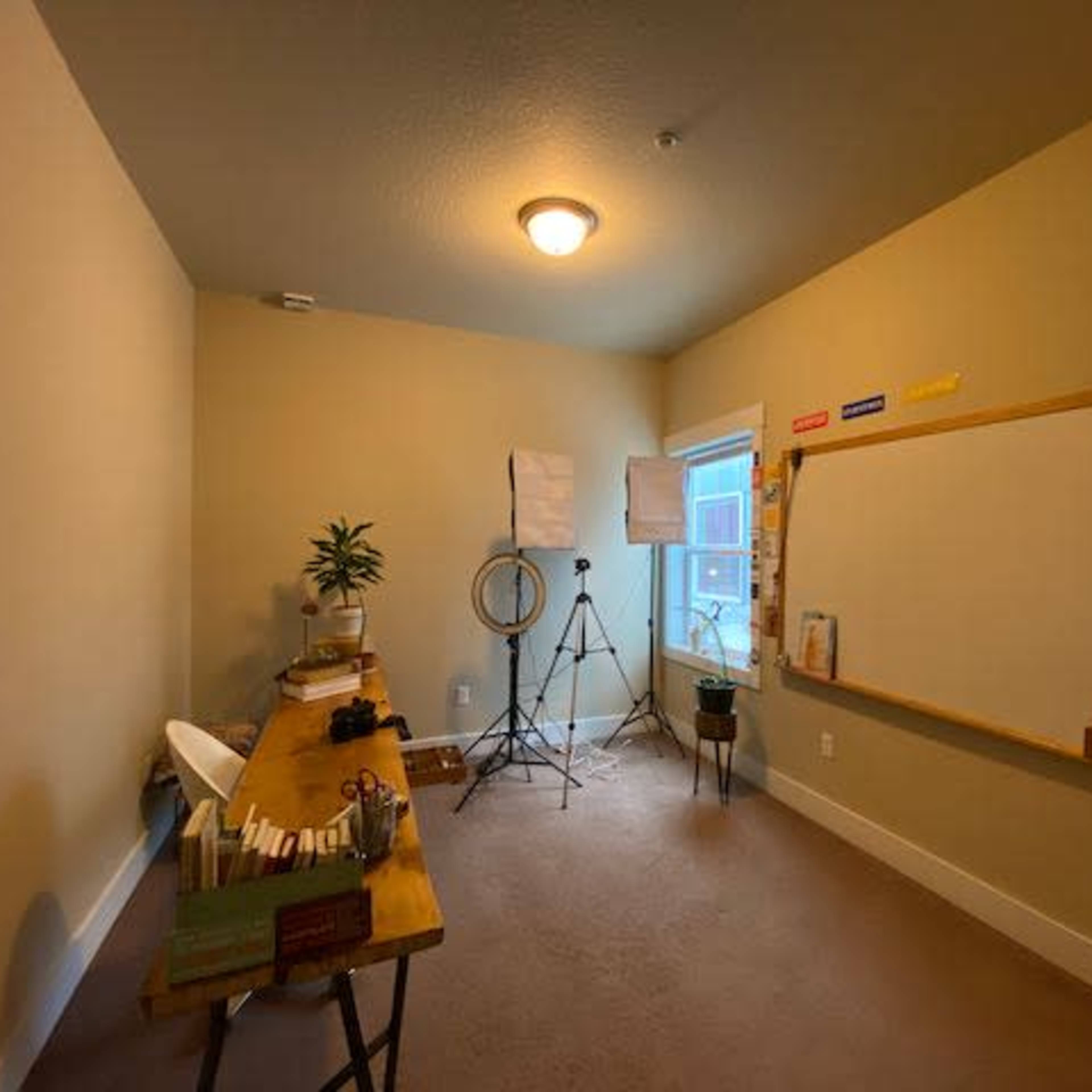 A small, well-lit room with a large whiteboard, a wooden desk with various items, and light equipment set up near a window.