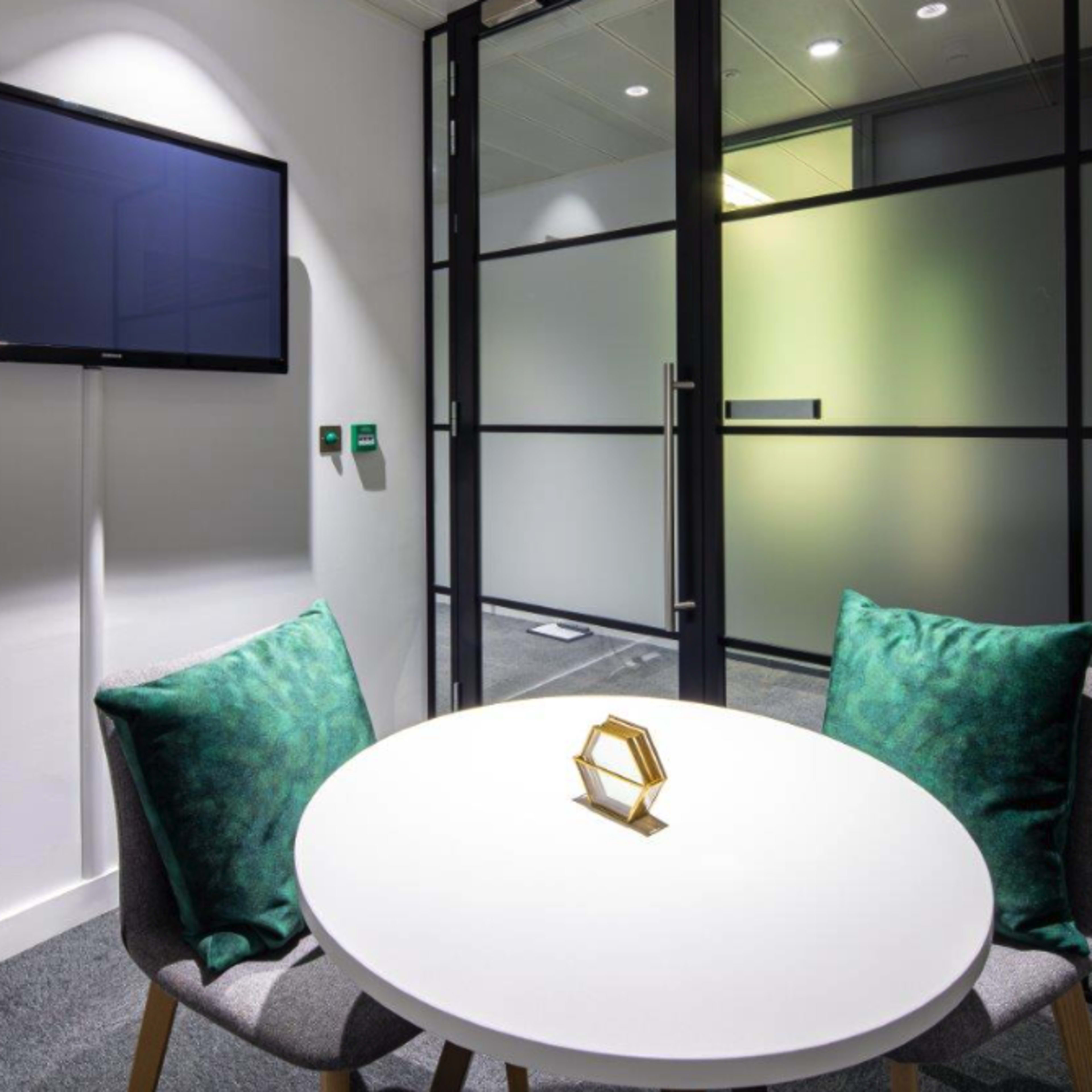 A modern meeting room features a round white table with two green-cushioned chairs and a wall-mounted television.