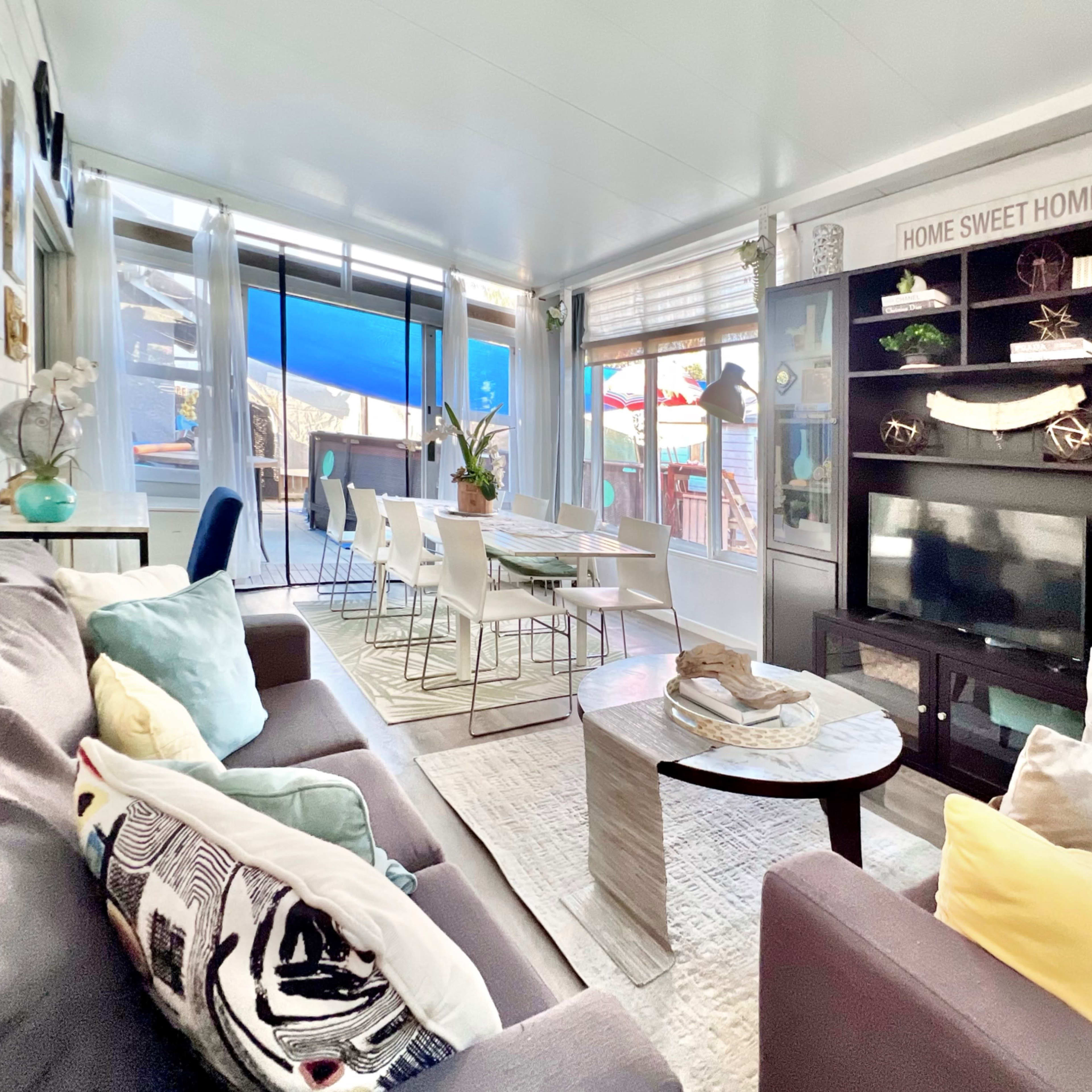 A bright living room features a comfortable couch, a central coffee table, a television stand with decorative items, and a dining area adjacent to large windows.