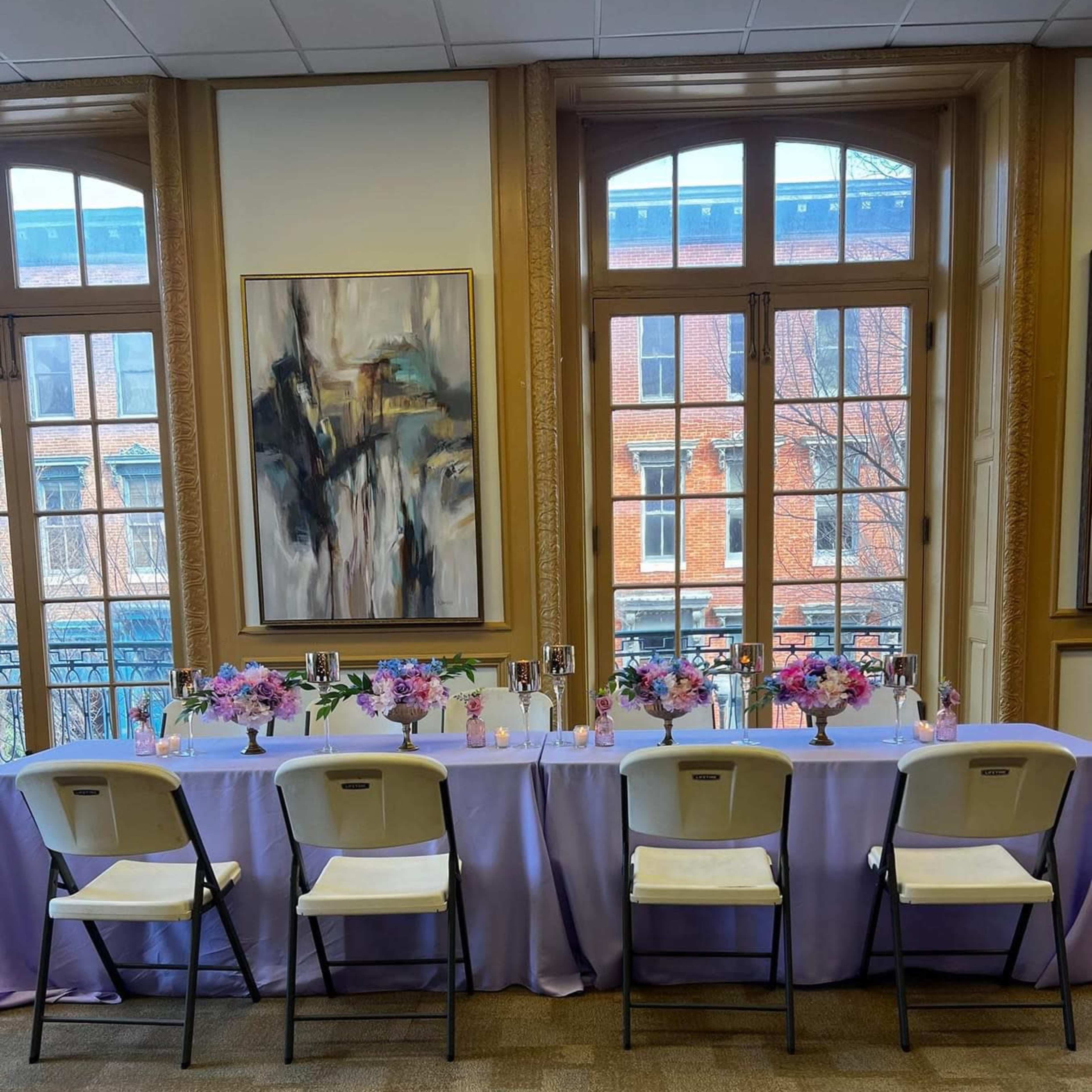 A long table is set with a lavender tablecloth and decorated with flower arrangements in vases, surrounded by white folding chairs and large windows that provide a view of a brick building outside.