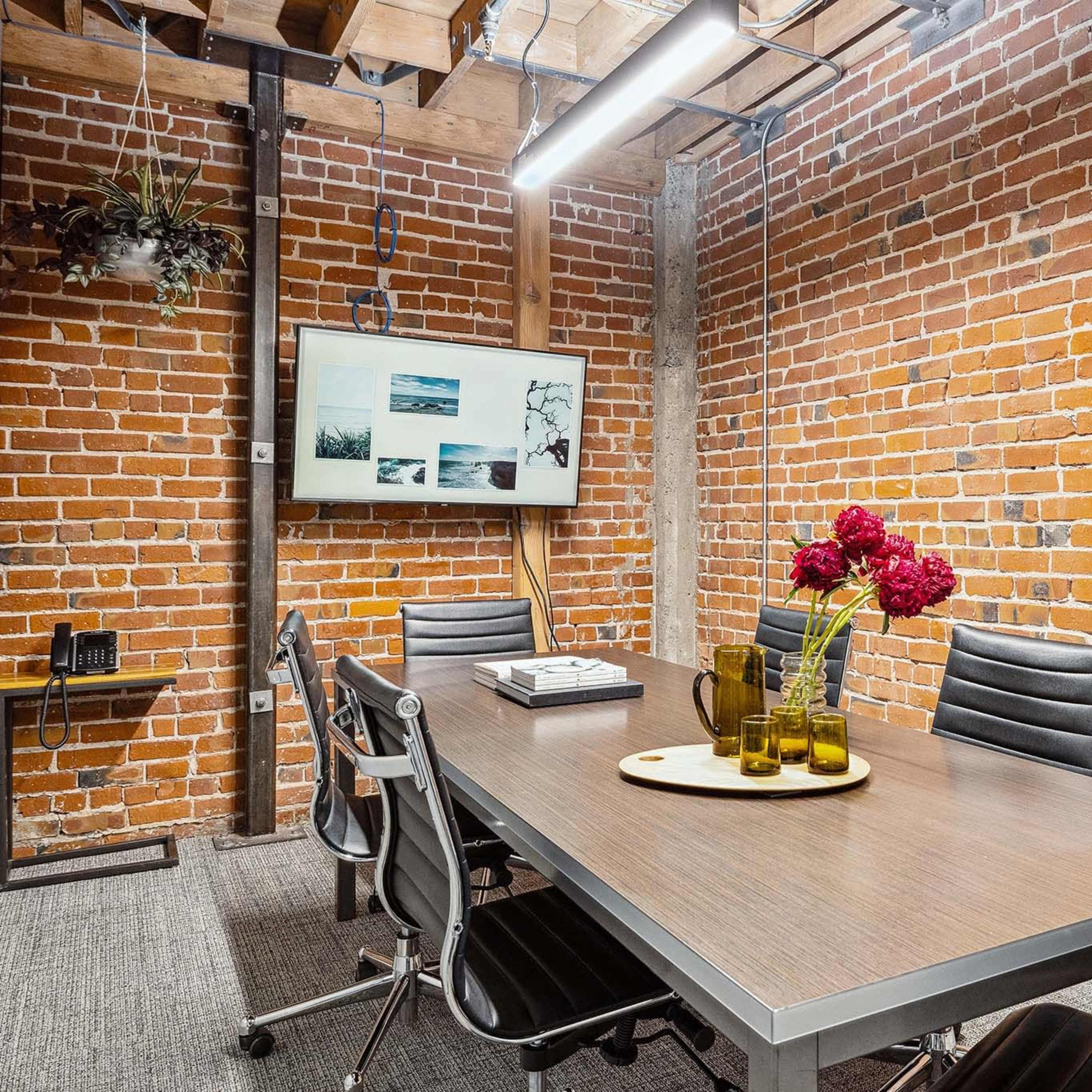 A conference room features a wooden table surrounded by black chairs, with exposed brick walls and a television displaying images.