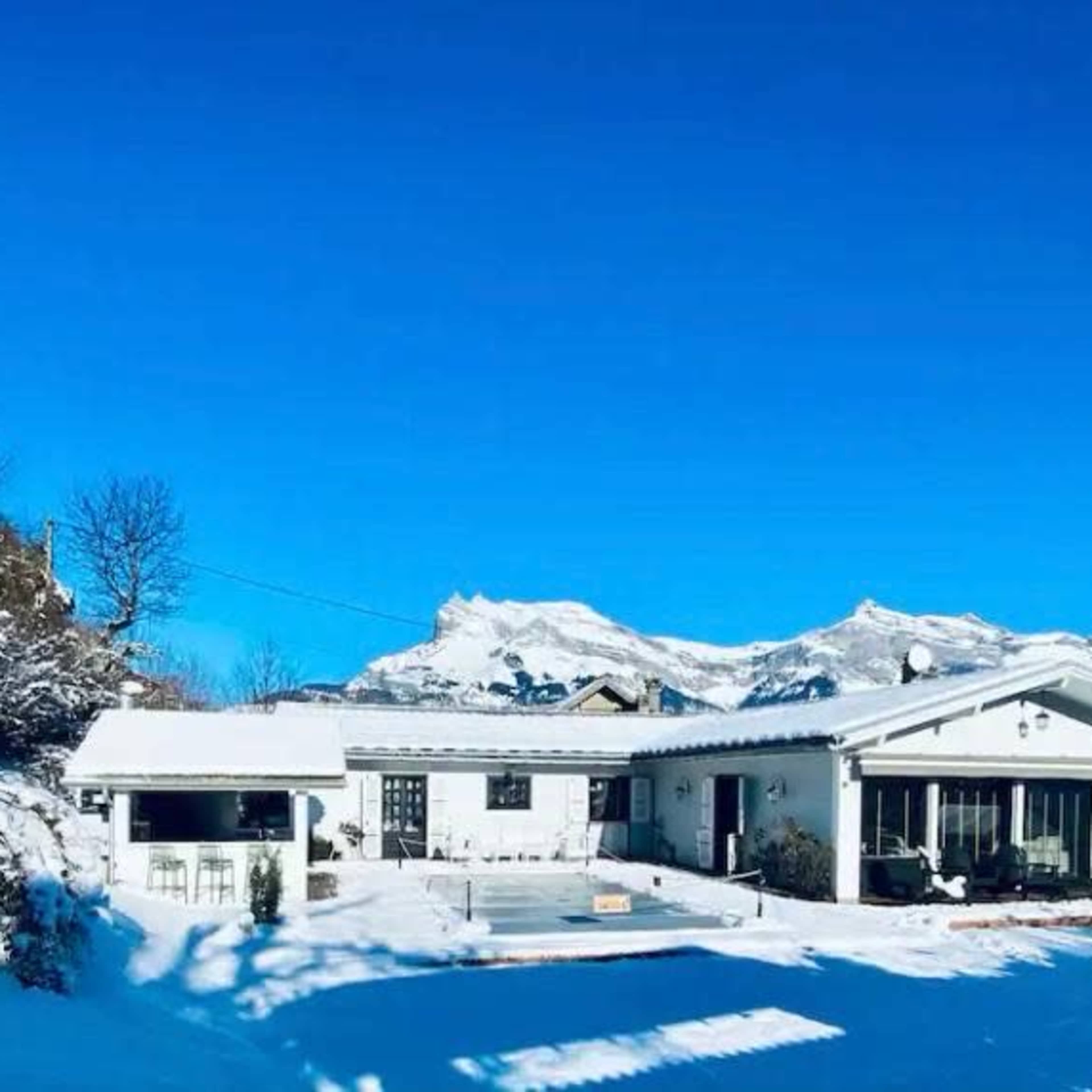 A snow-covered house sits against a backdrop of mountains under a clear blue sky.