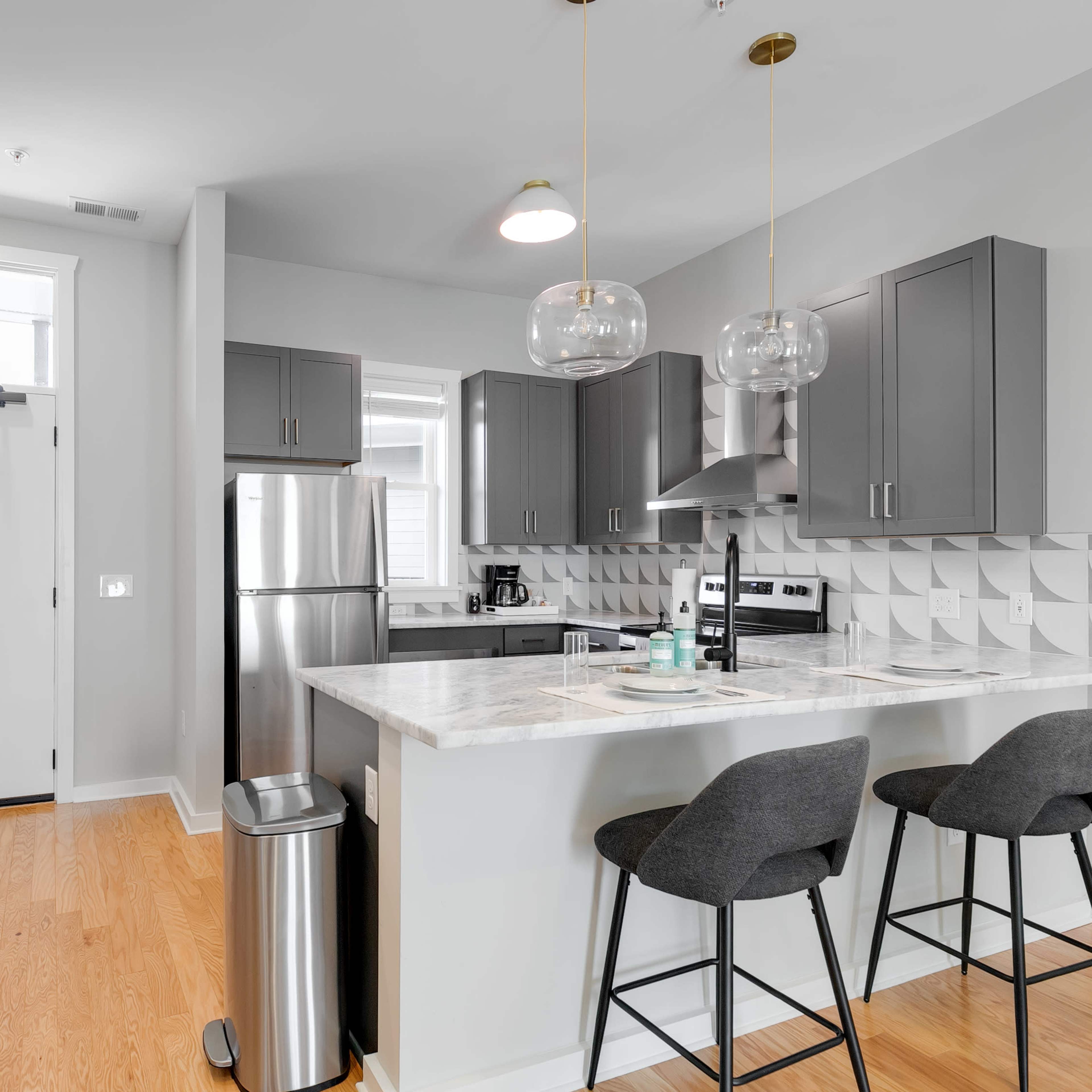 A modern kitchen with gray cabinetry, stainless steel appliances, and a breakfast bar with two stools, adjacent to a front door.