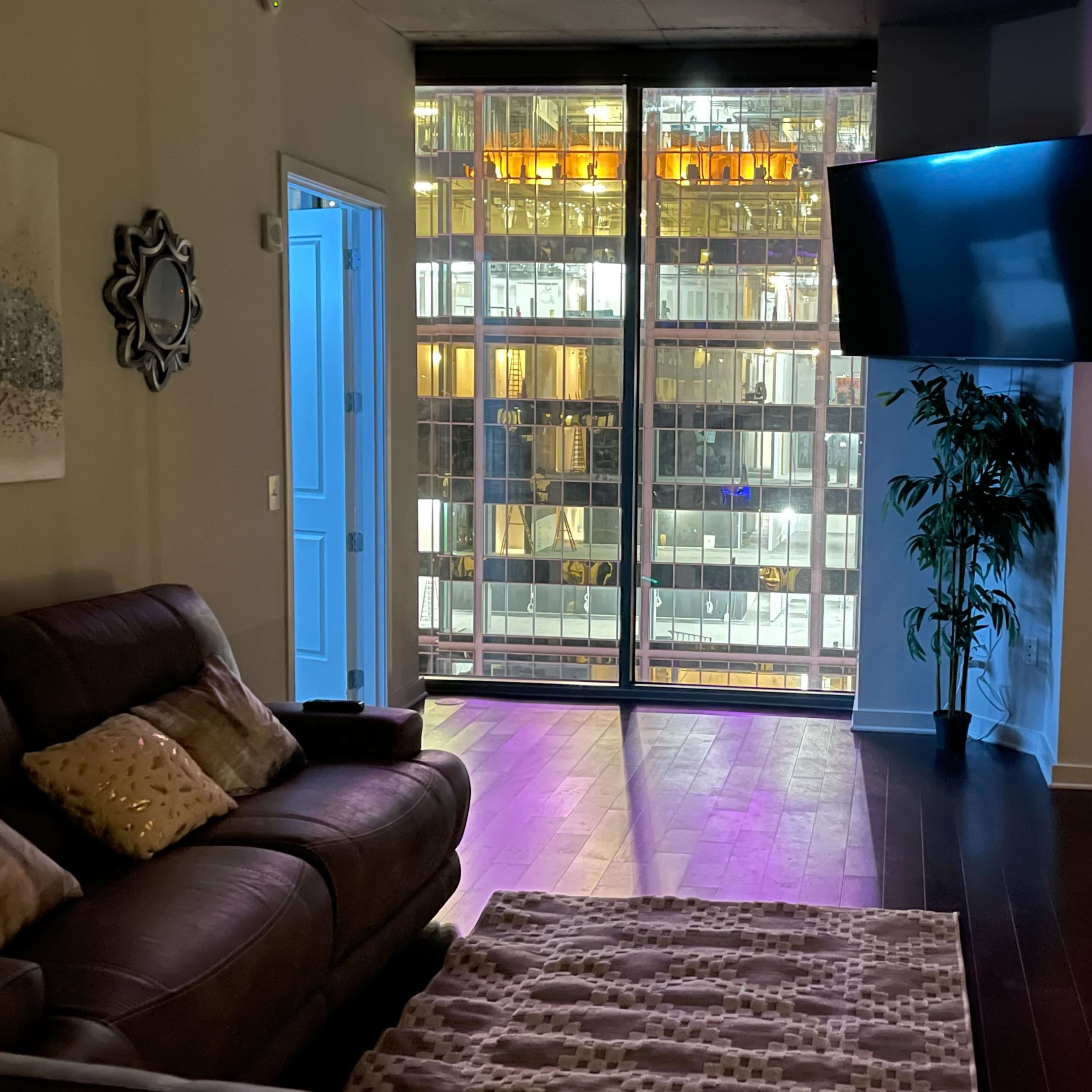 The living room features a brown leather sofa, a television mounted on the wall, and large windows showcasing a city skyline at night.