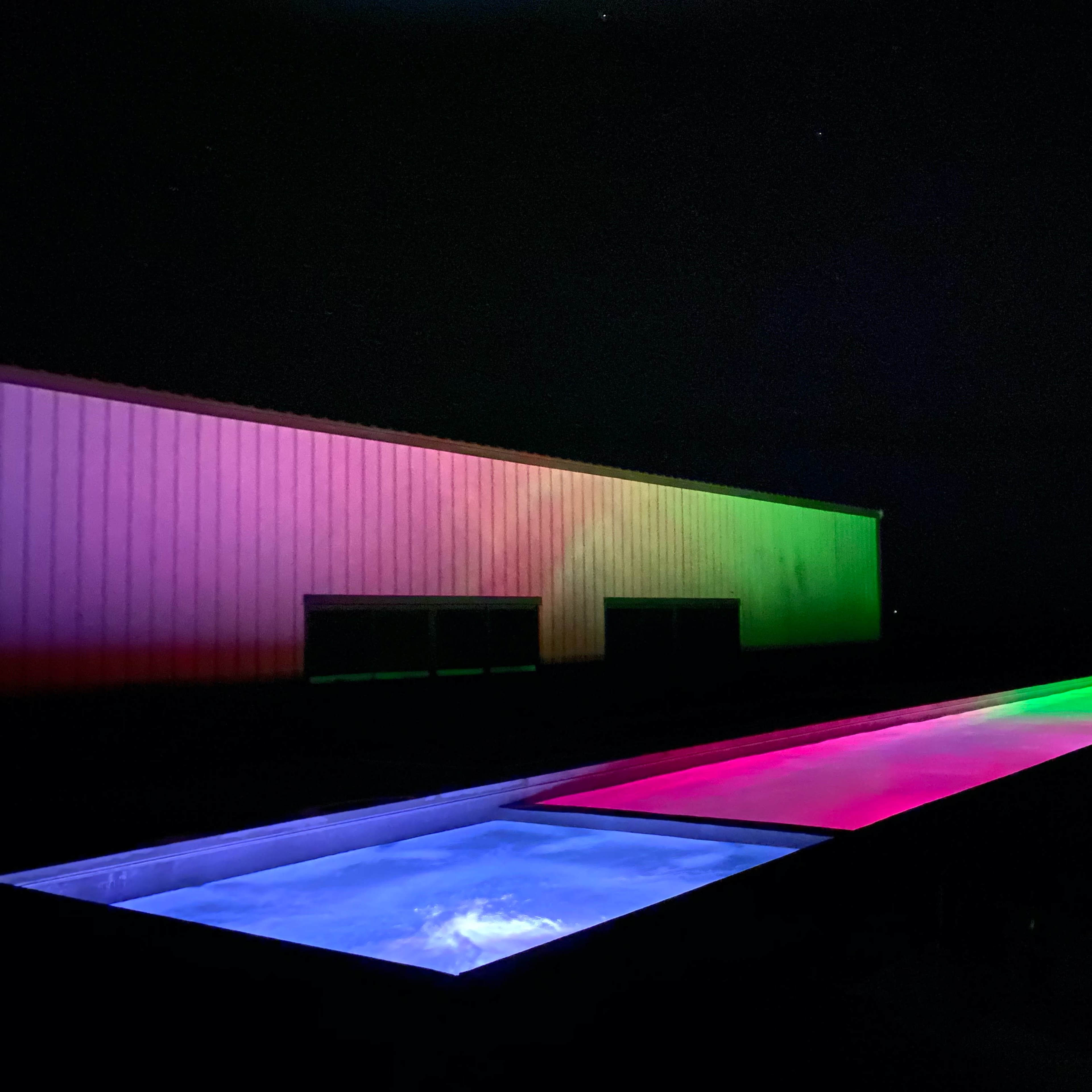 A long, illuminated outdoor pool is flanked by a building with colorful lighting in the background at night.