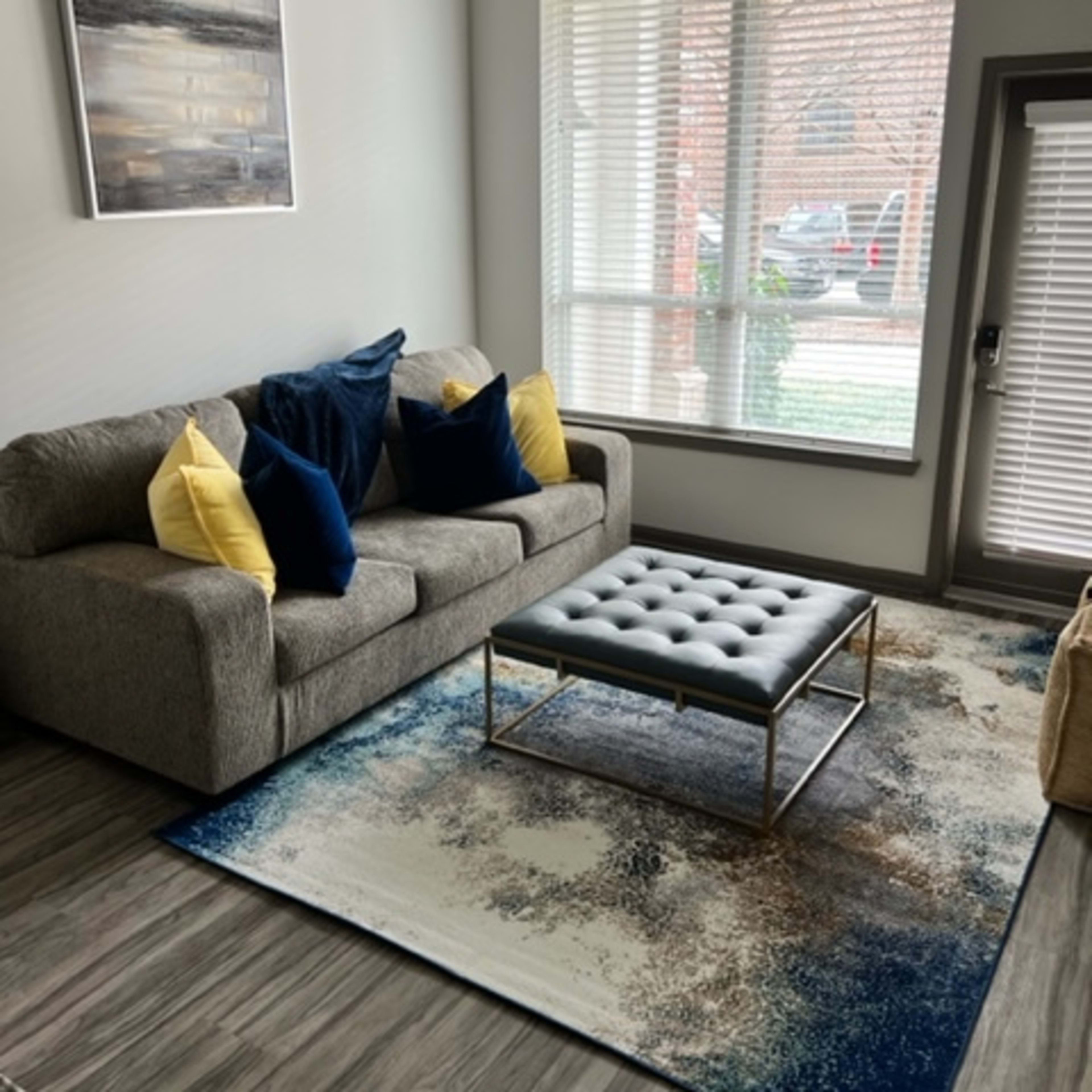 A living room with a grey sofa, two yellow and blue pillows, a tufted ottoman on a patterned rug, and large windows allowing natural light.