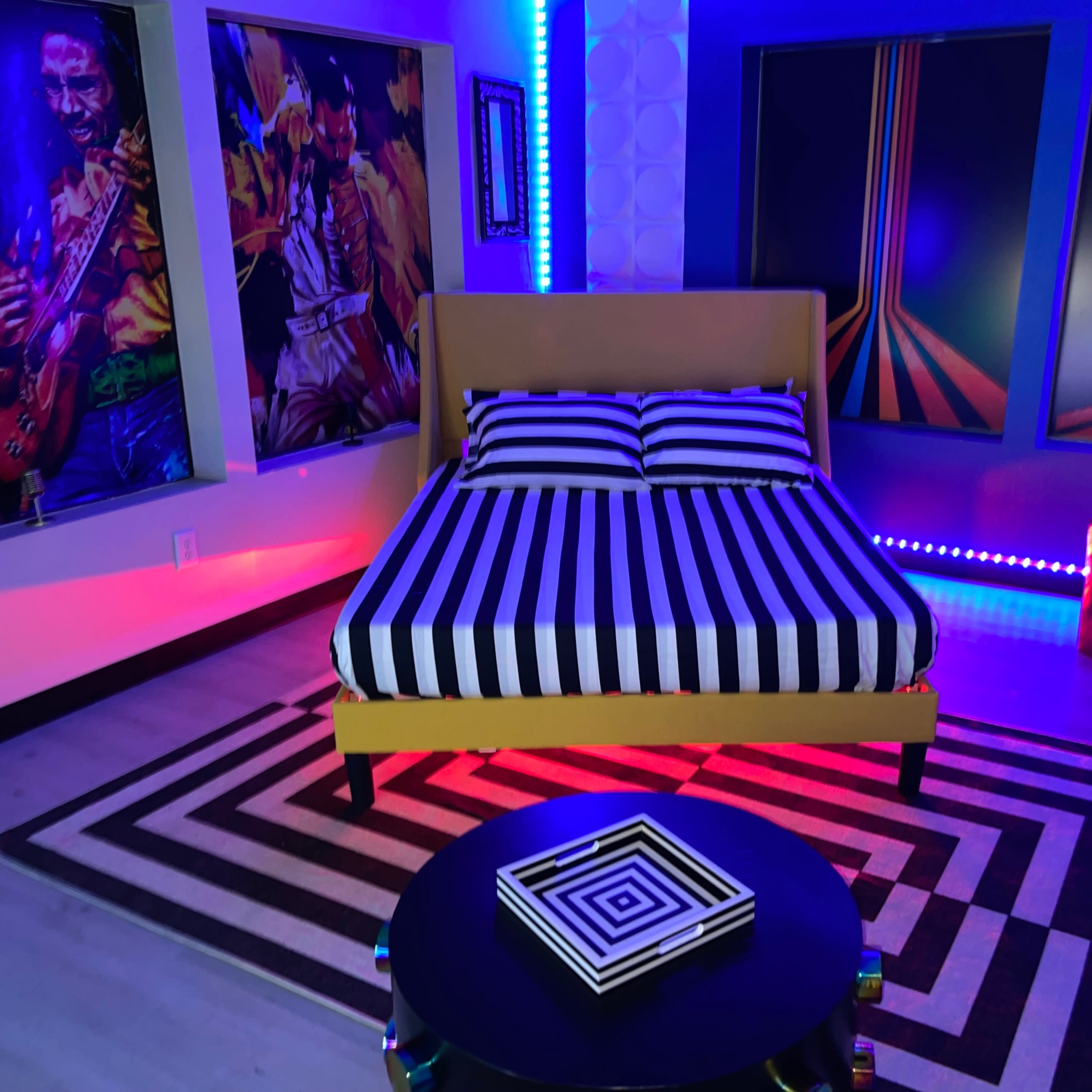 The scene depicts a bedroom with a yellow bed featuring black and white striped bedding, surrounded by colorful wall artwork and vibrant LED lights.