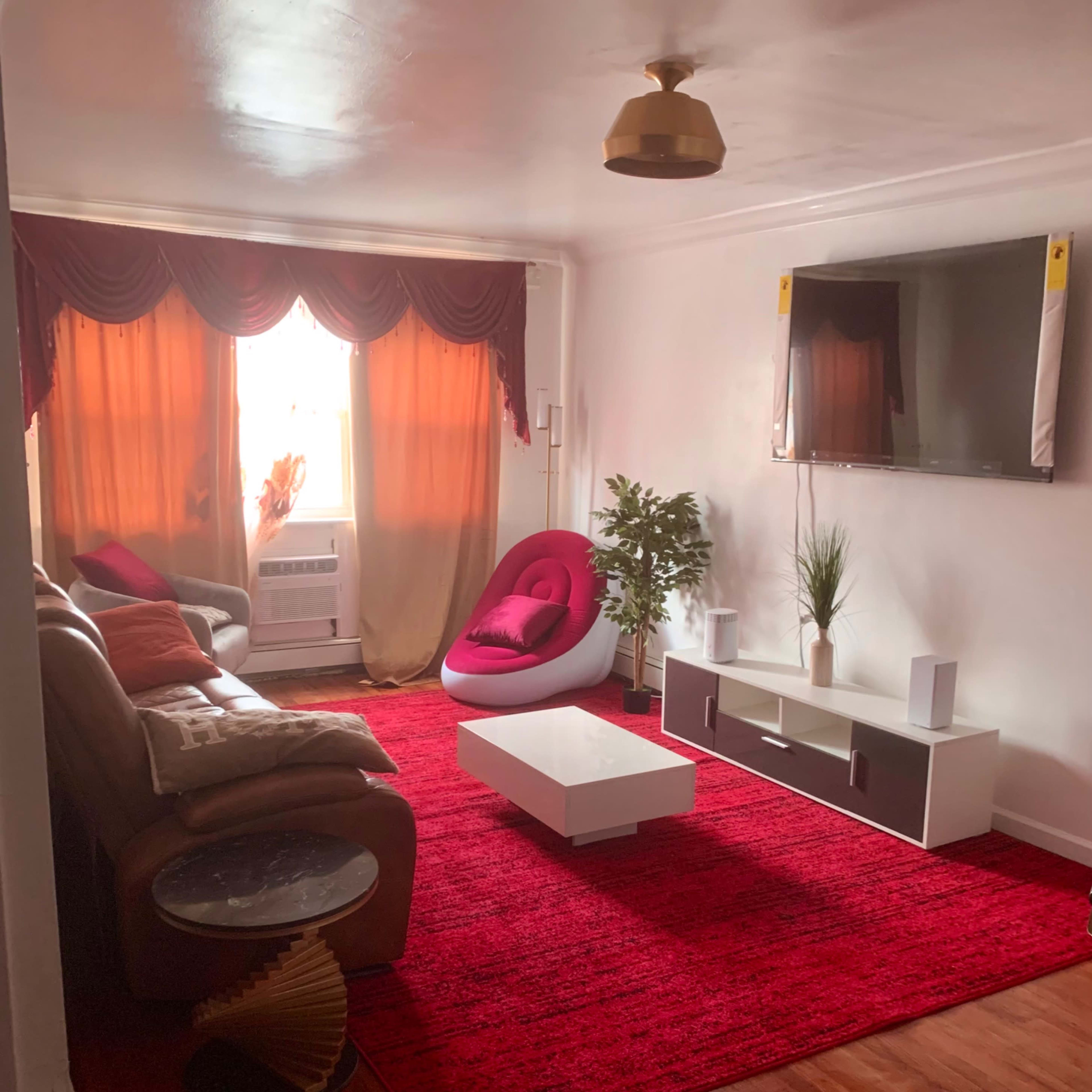 The living room features a red area rug, a white television stand, a large window with curtains, and a mix of seating including a brown couch and a modern white chair.