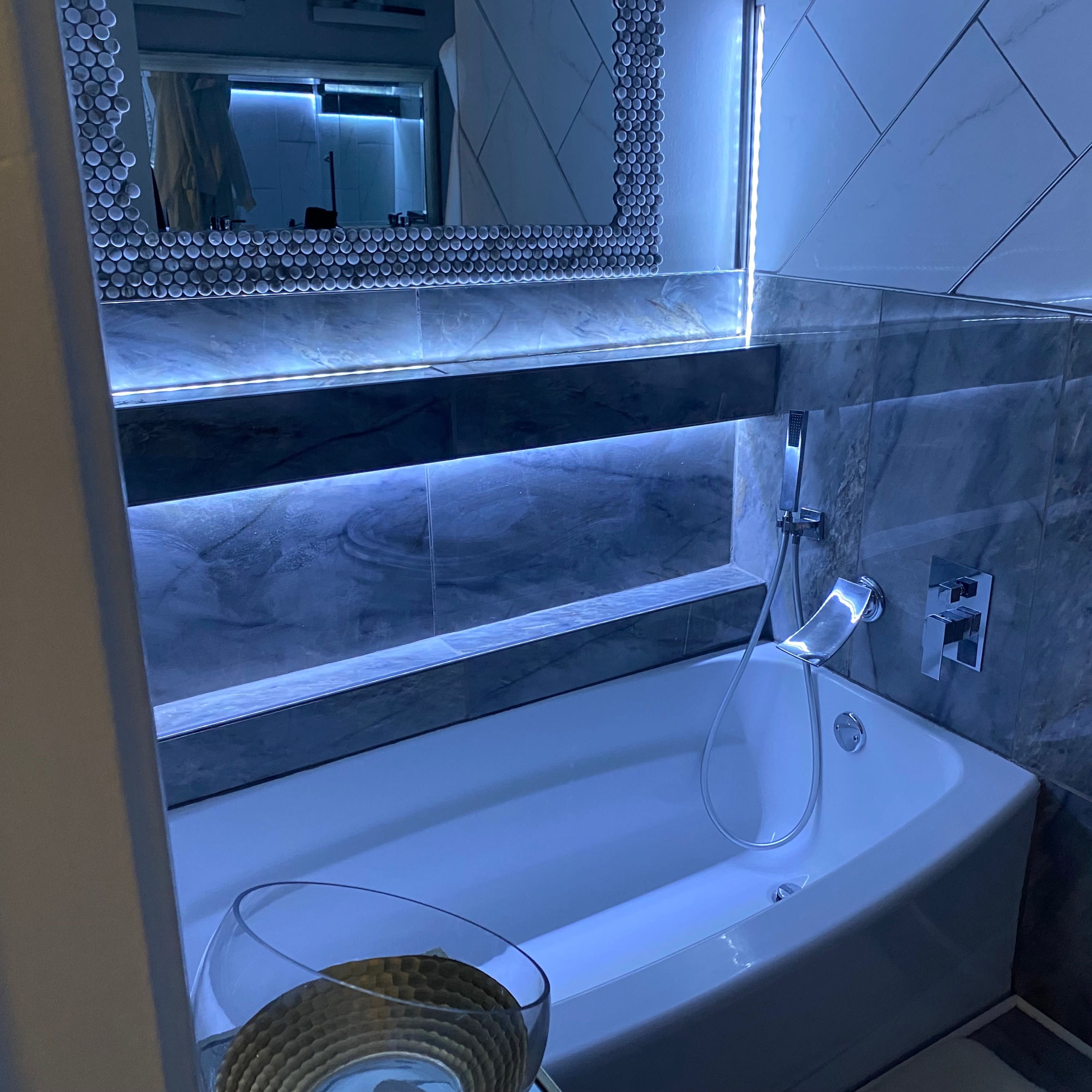 A modern bathroom with a curved bathtub, a large mirror, and ambient lighting along the tiled walls.