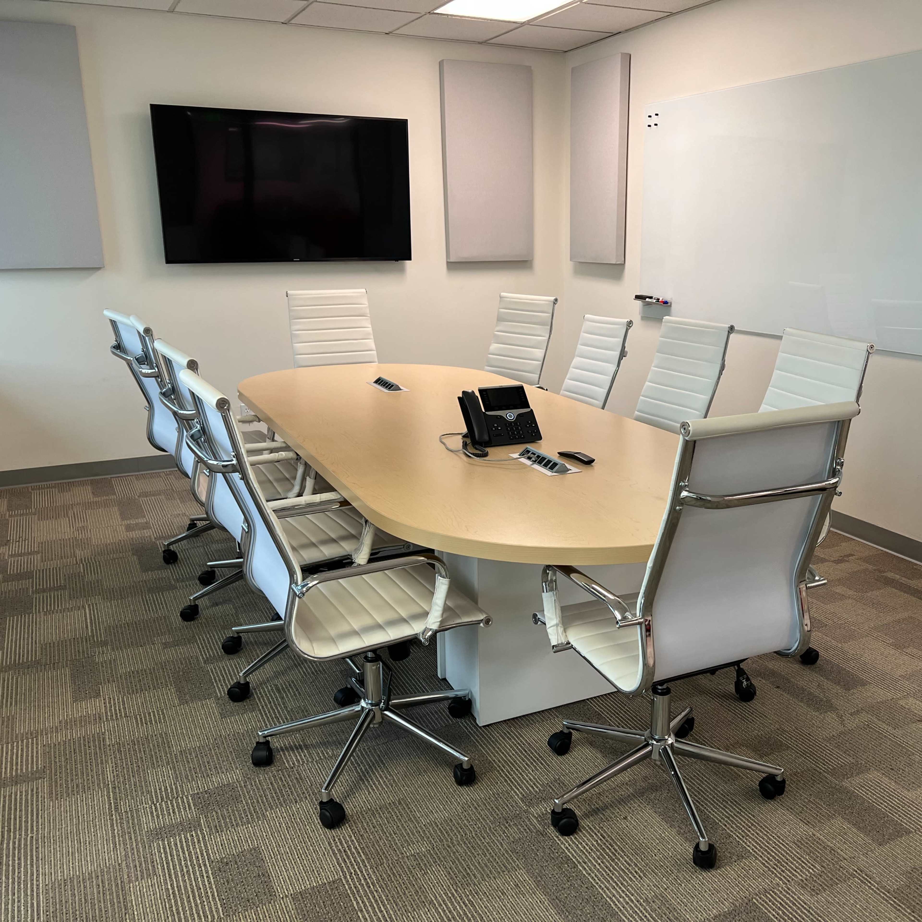 A modern conference room is set up with an oval table surrounded by white ergonomic chairs, a wall-mounted screen, and a whiteboard.