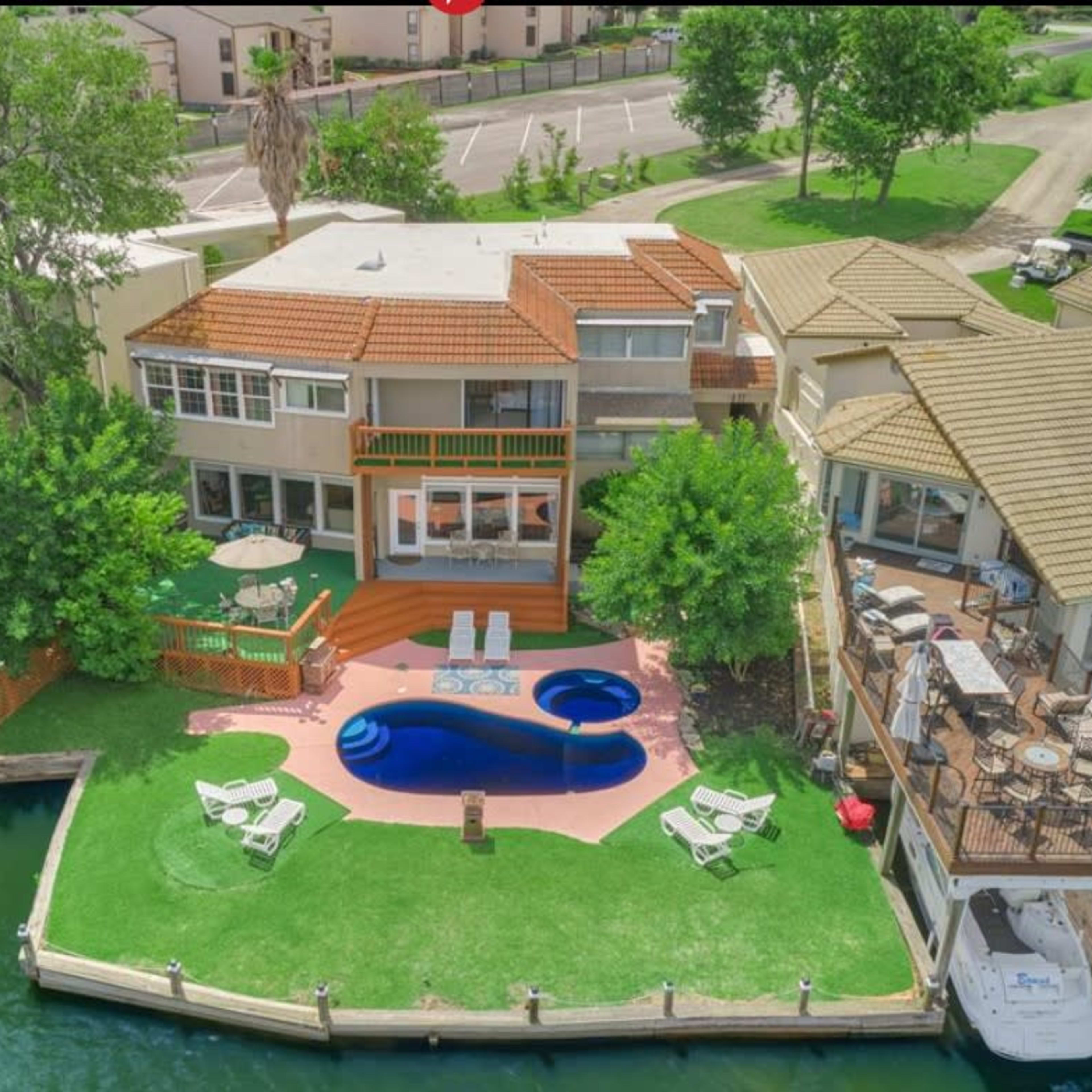 A large waterfront property with a swimming pool, multiple decks, and boats docked on a green lawn alongside a canal.