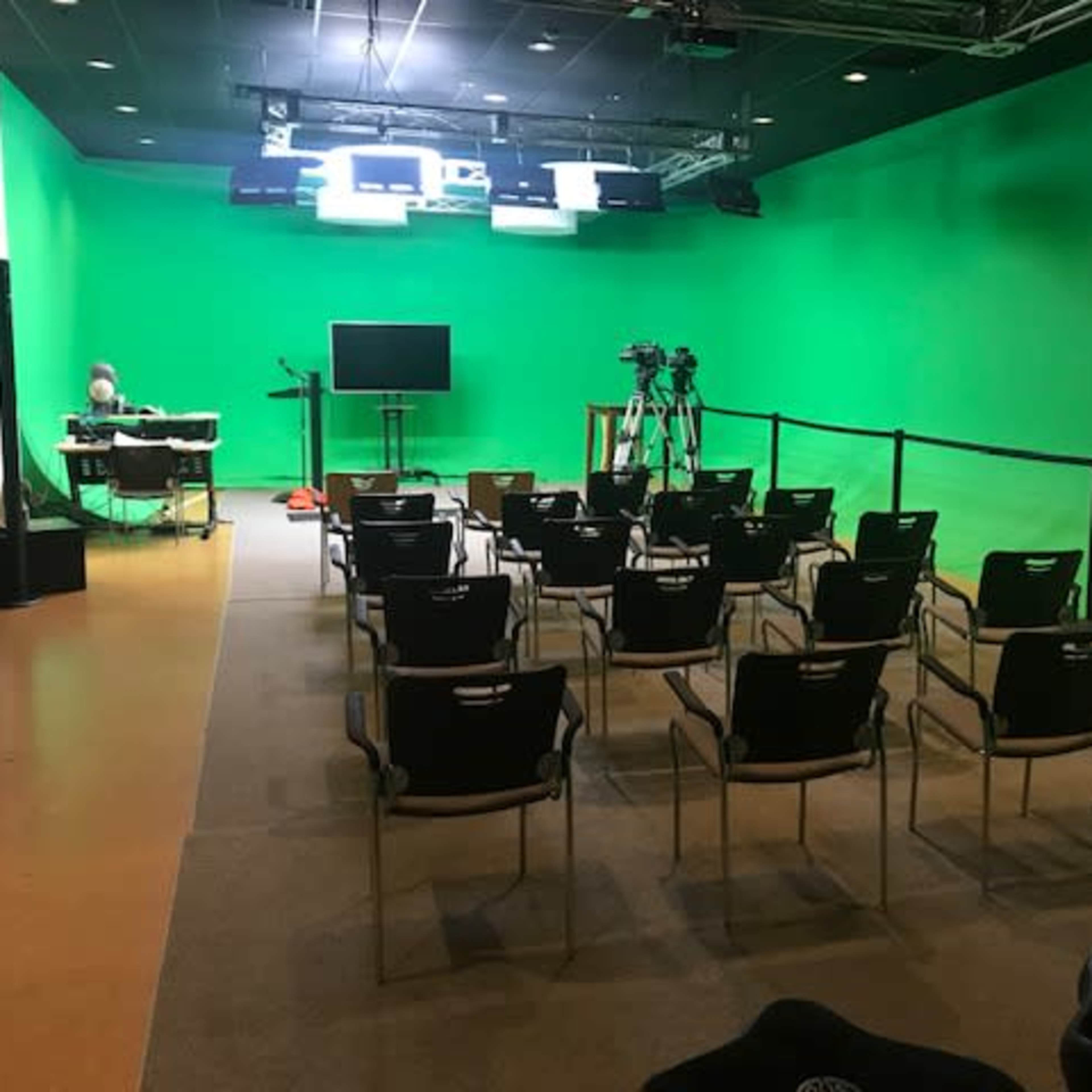 A green screen studio with rows of empty chairs, a table with equipment, and cameras set up for filming.