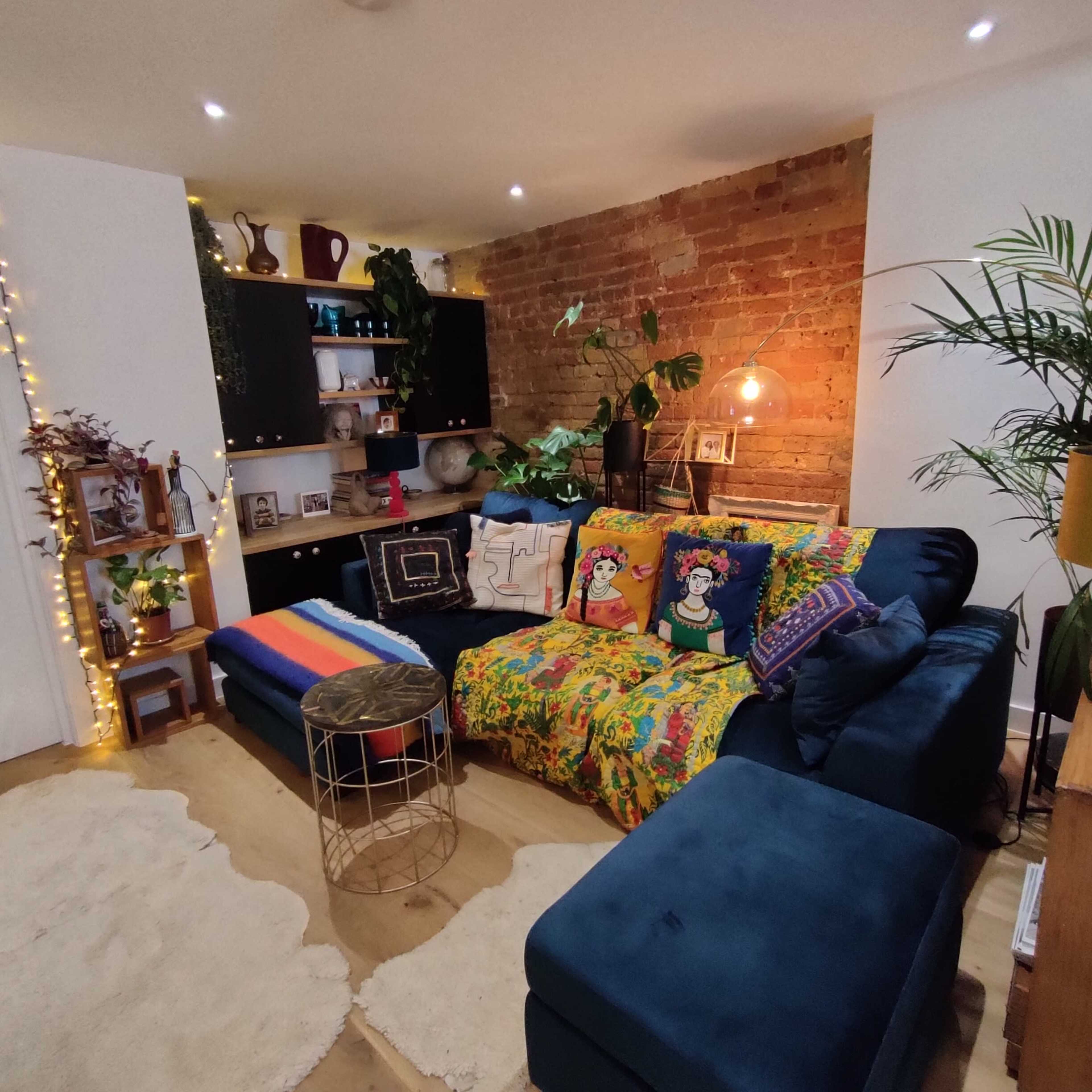 A cozy living room features a blue sectional sofa adorned with colorful pillows and a vibrant throw, set against a backdrop of exposed brick walls and a shelf displaying decorative items.