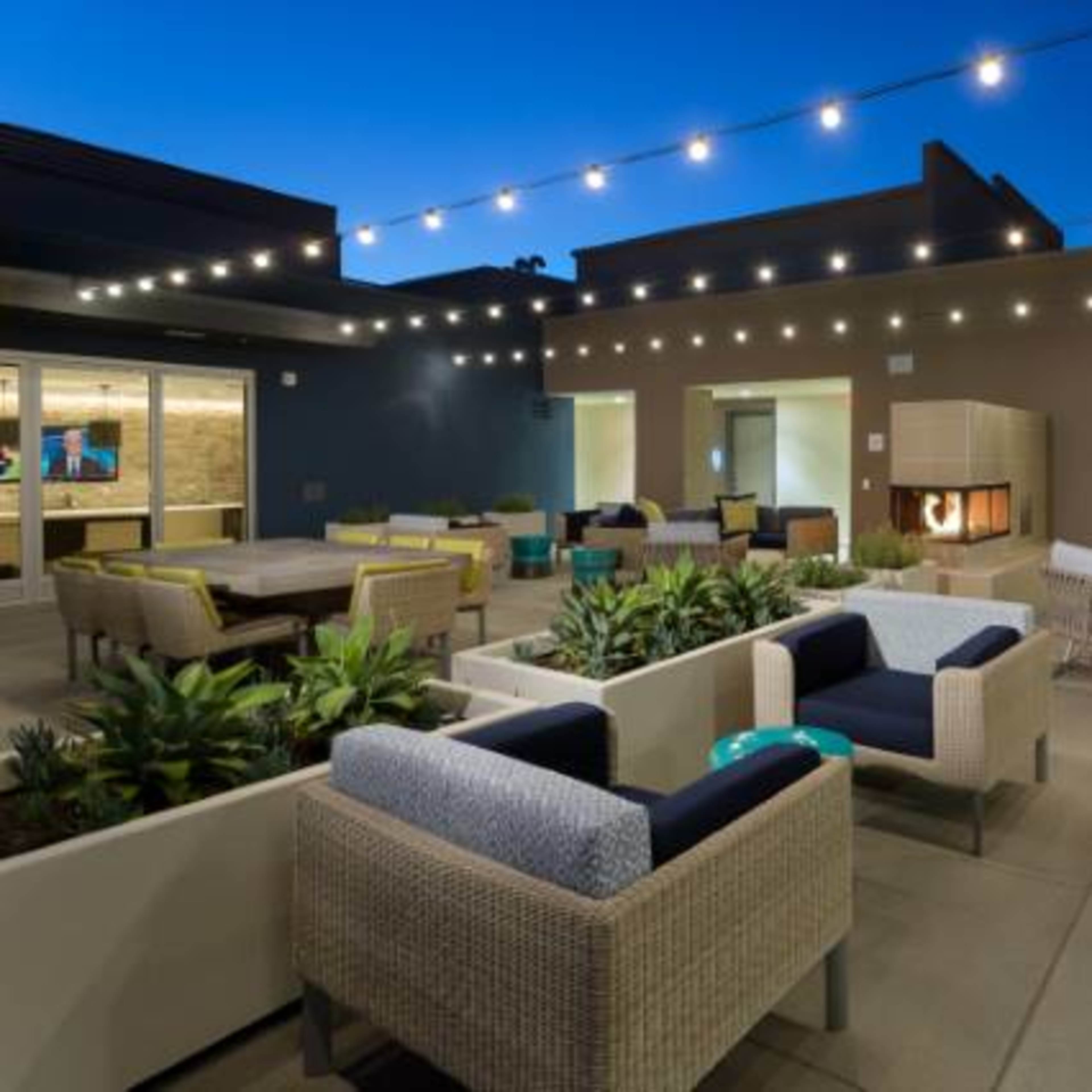 A modern outdoor lounge area with seating, fire features, and string lights illuminating the space at dusk.