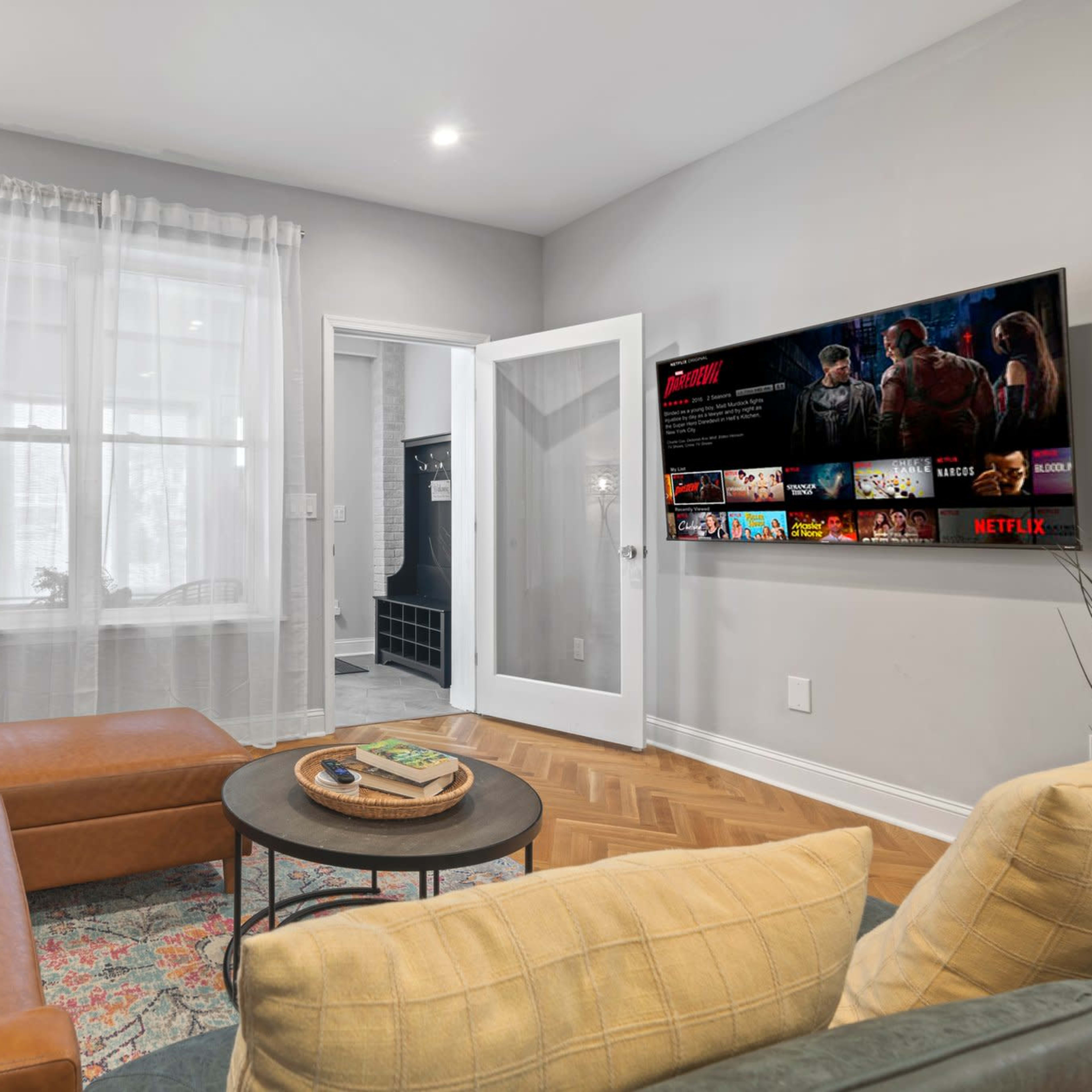 A cozy living room features a sectional sofa, a round coffee table, and a wall-mounted TV displaying a streaming service.
