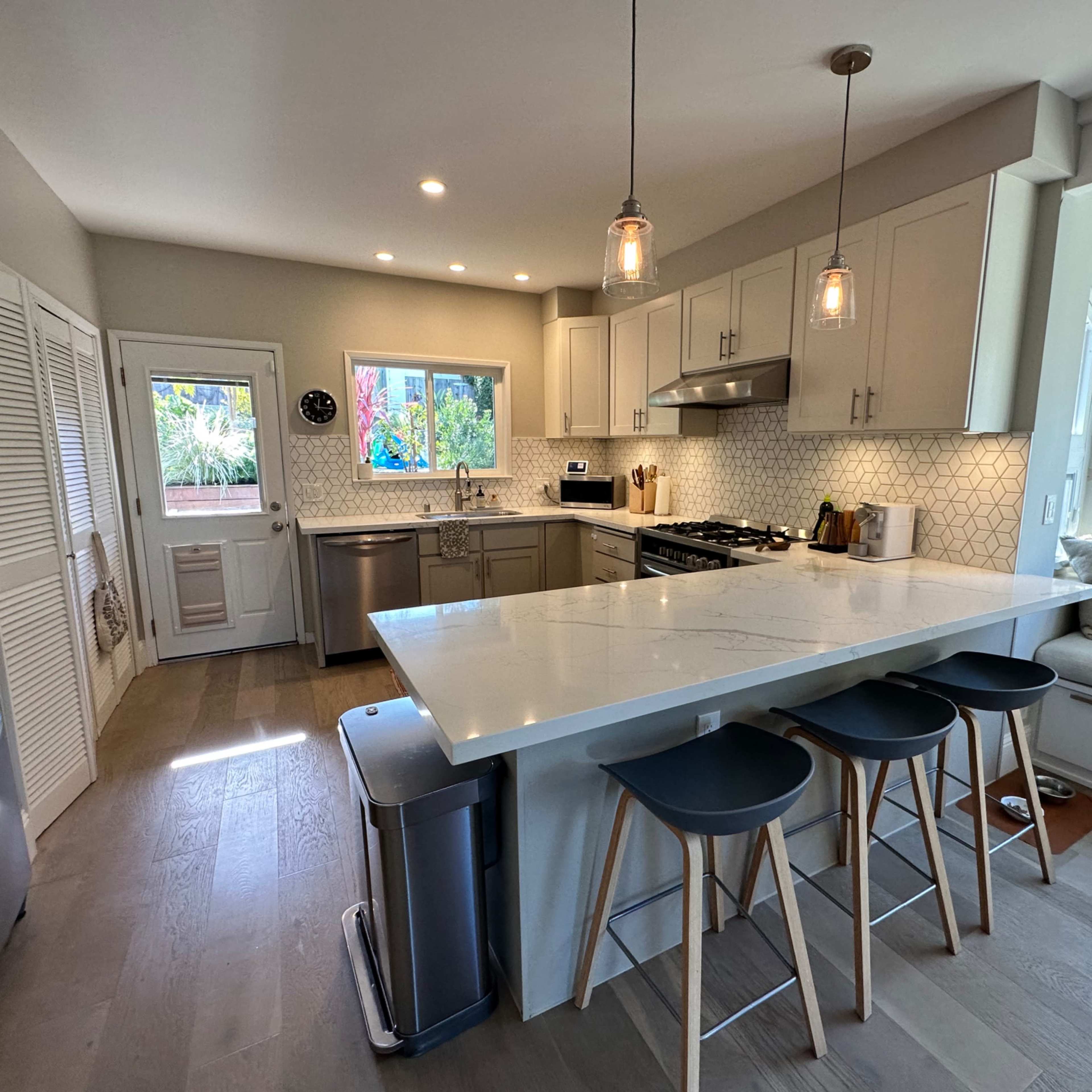 A modern kitchen features a large island with three stools, stainless steel appliances, and ample natural light from large windows.