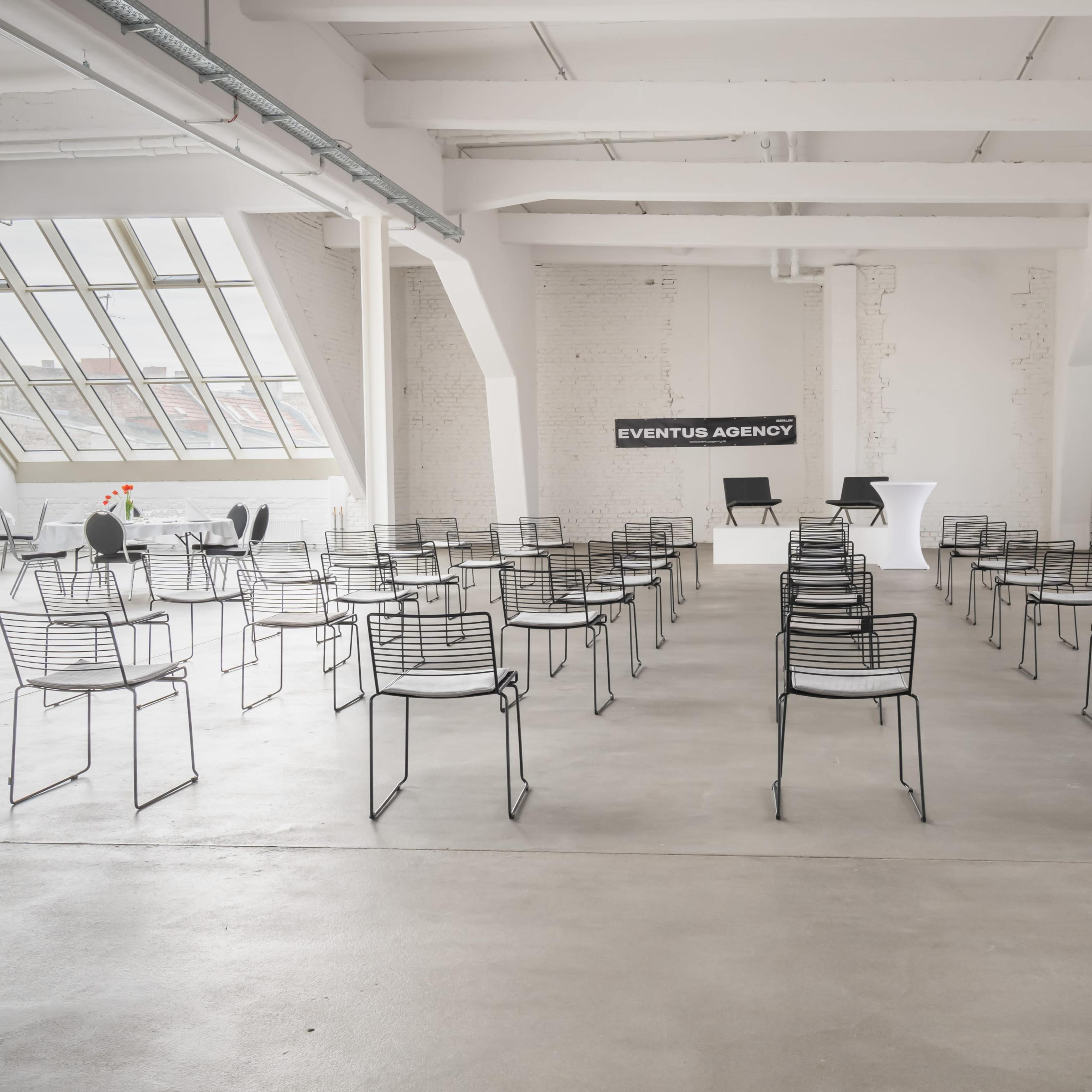A spacious, minimalist conference room with rows of empty black wire chairs arranged for an event, featuring a backdrop that reads "EVENTUS AGENCY."