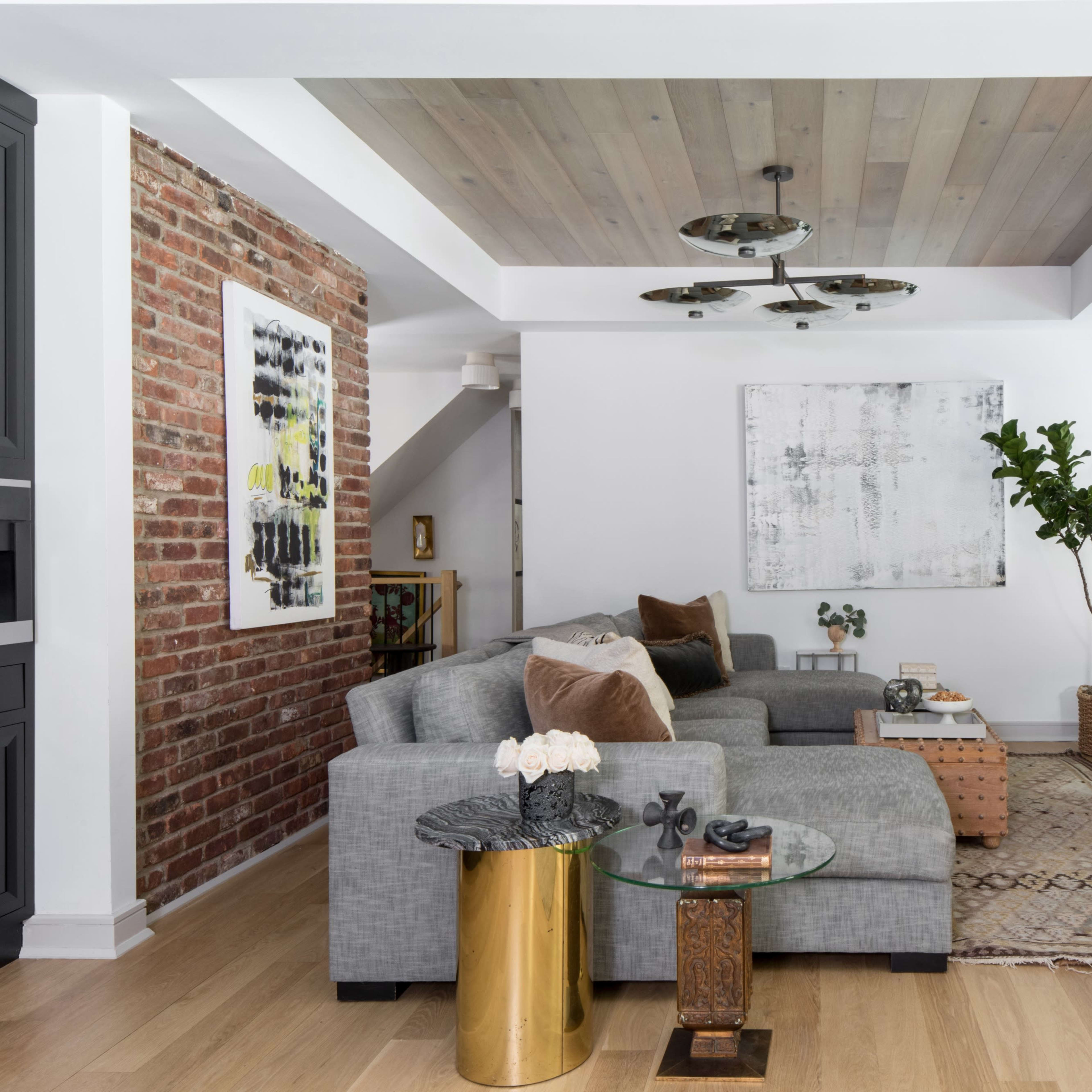 A modern living room featuring a gray sectional sofa, a coffee table, a wall-mounted TV, and a lush green plant, situated beside exposed brick and wooden ceiling elements.