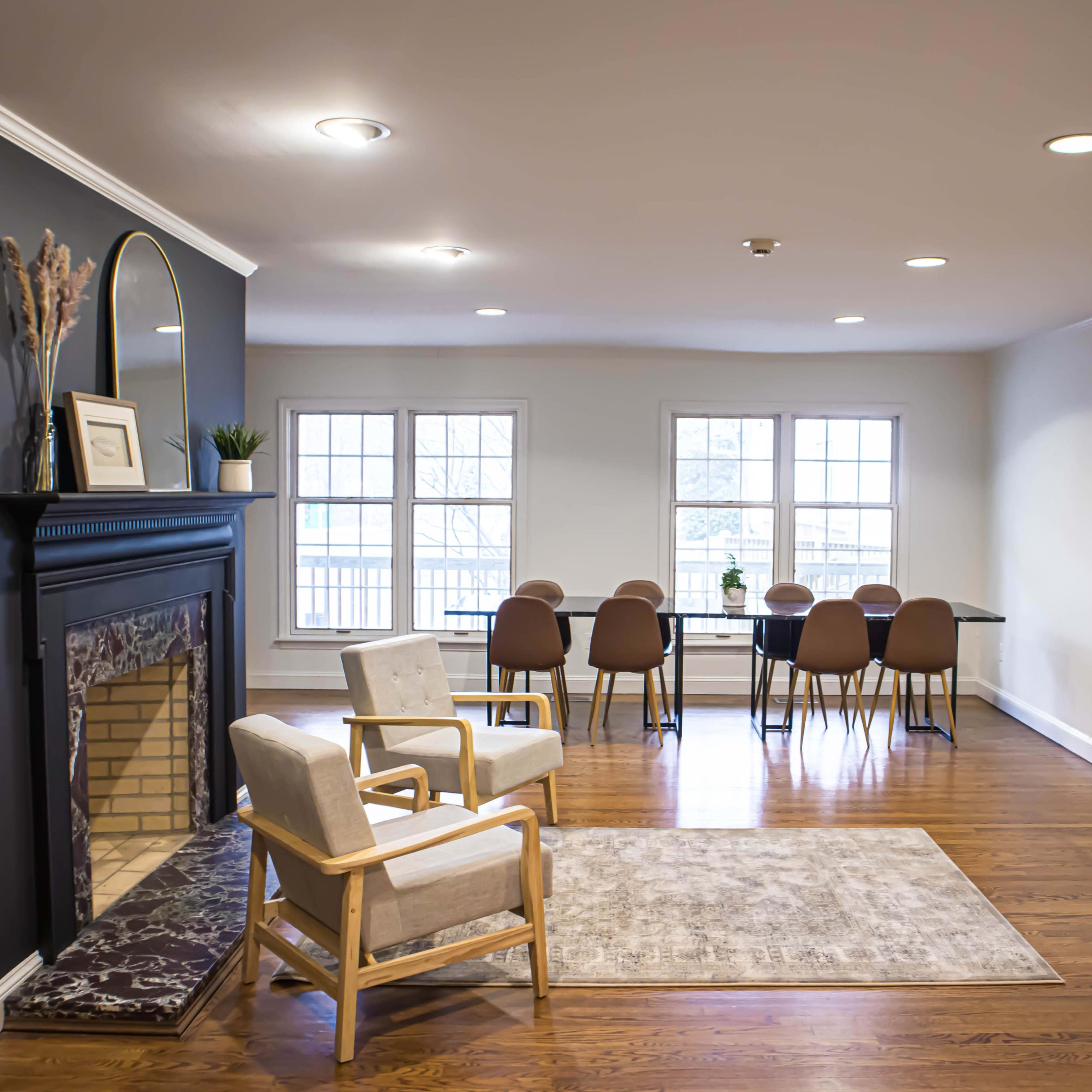 A spacious living area featuring a white chair and a fireplace on one side, with a dining table and chairs set up in front of large windows on the opposite side.
