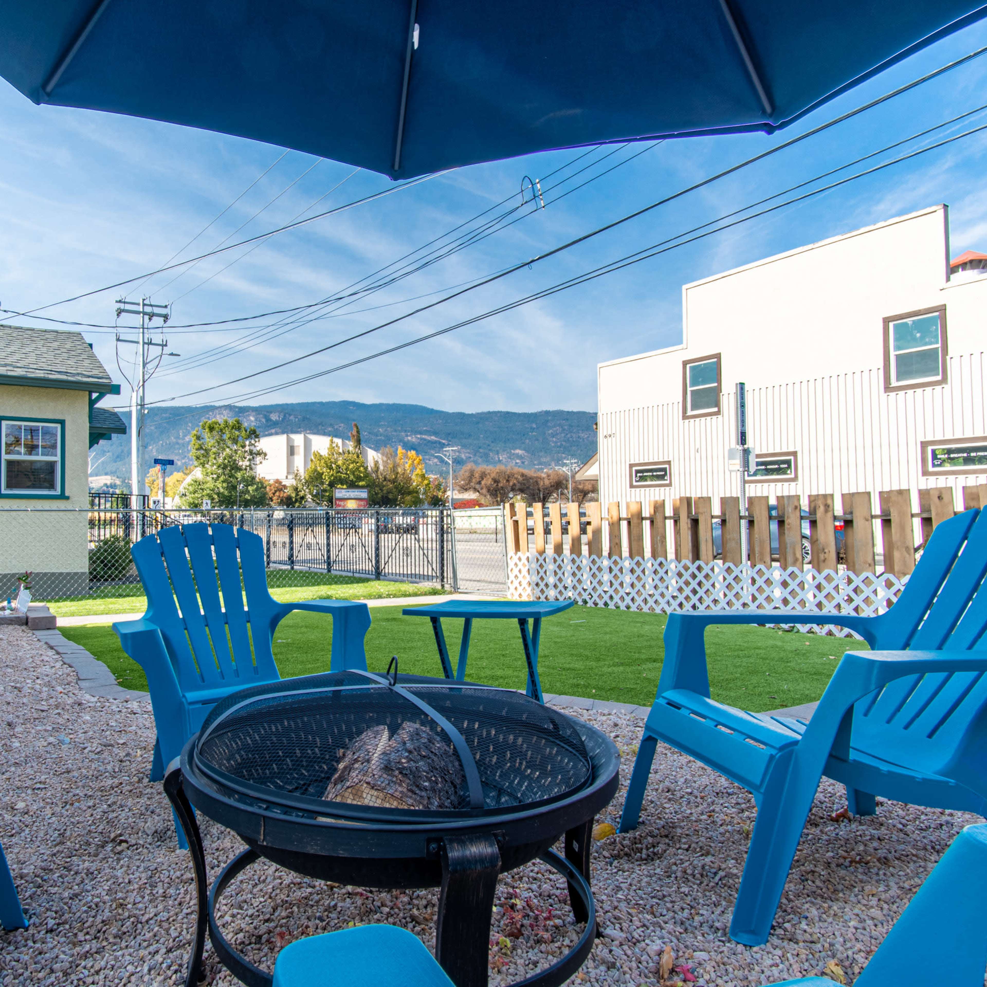 A gravel yard features a fire pit surrounded by blue Adirondack chairs, with a backdrop of mountains and buildings under a clear sky.