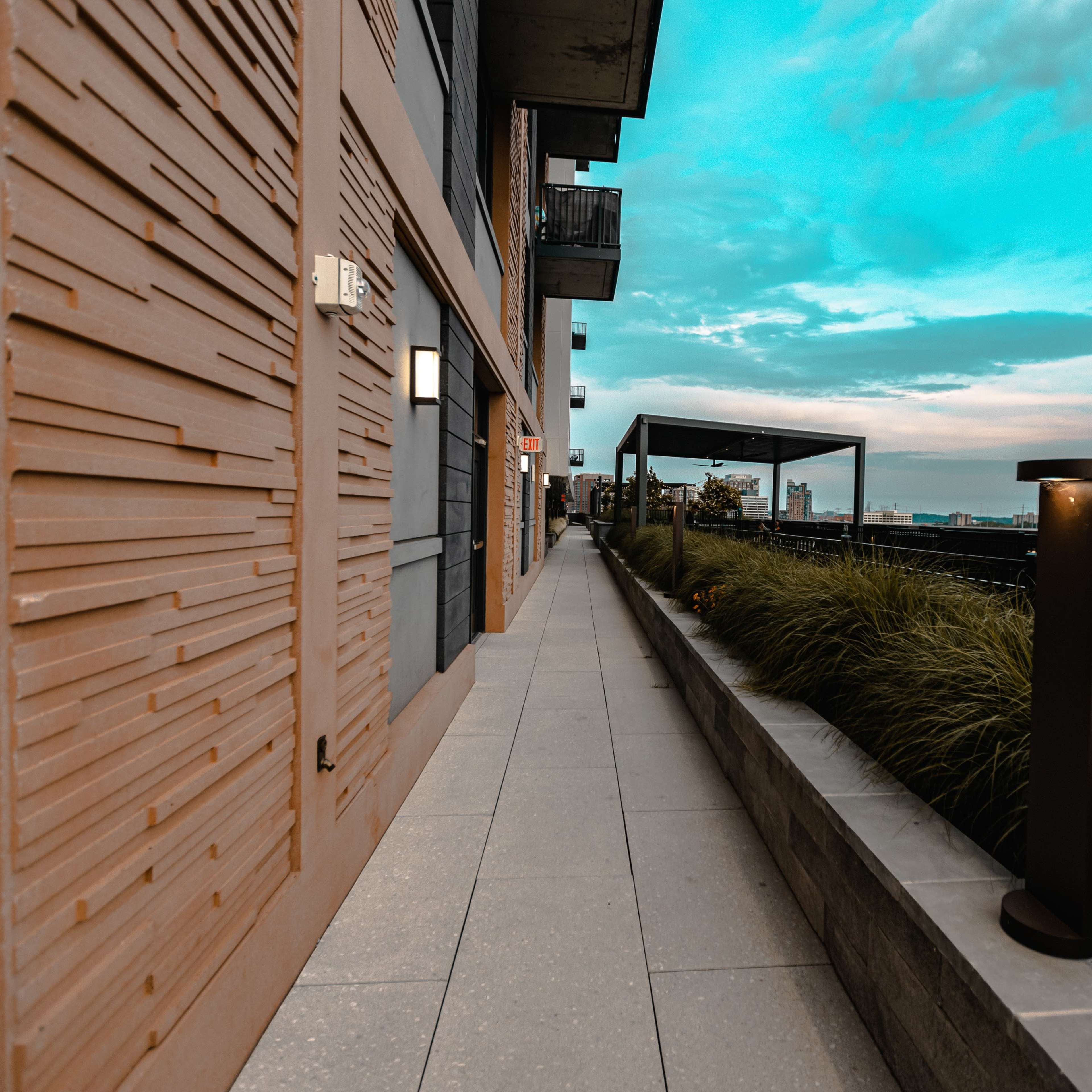 A narrow pathway lined with grass and modern lighting runs alongside a textured building under a blue sky.