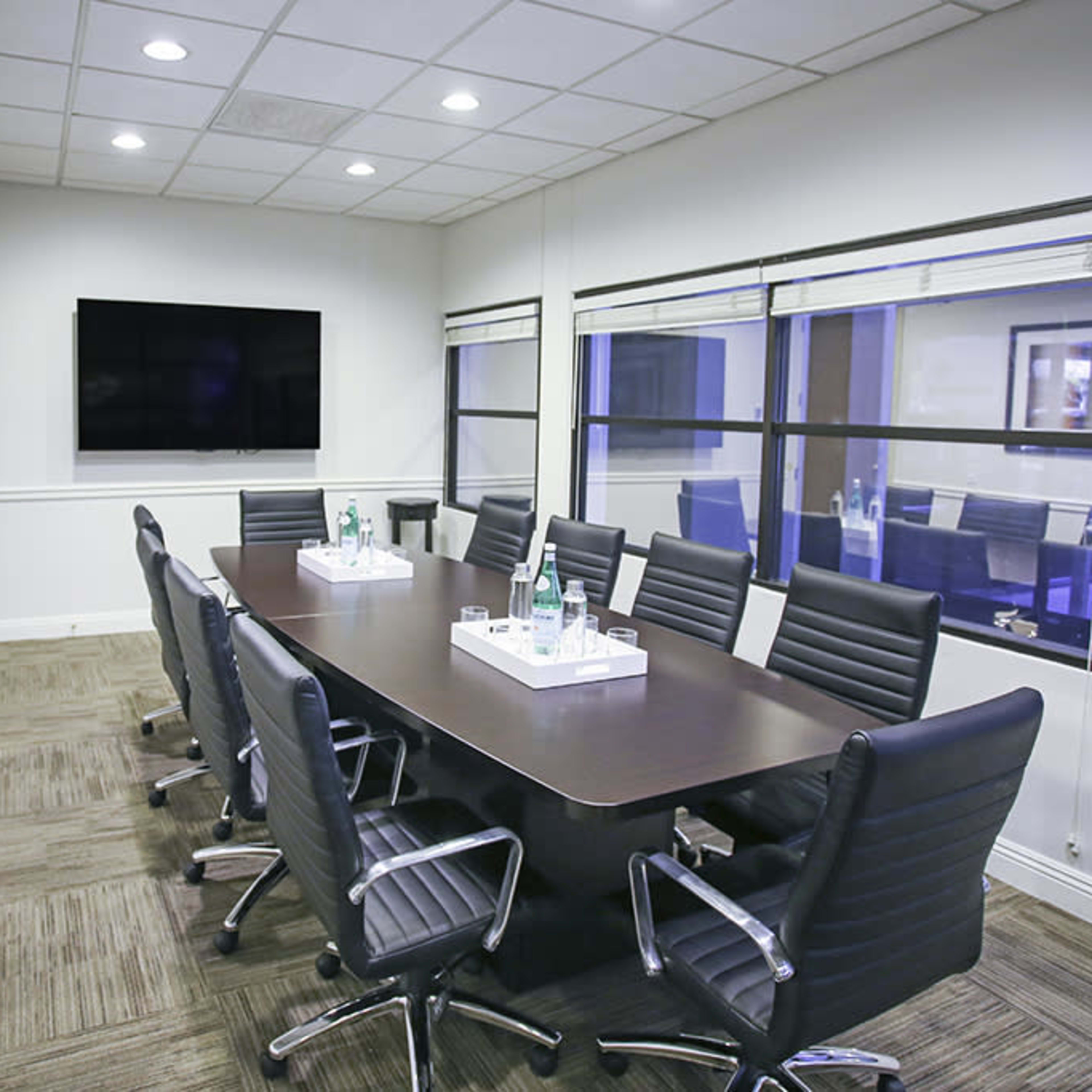 A conference room features a long wooden table surrounded by black chairs, with a television mounted on the wall and large windows allowing natural light.