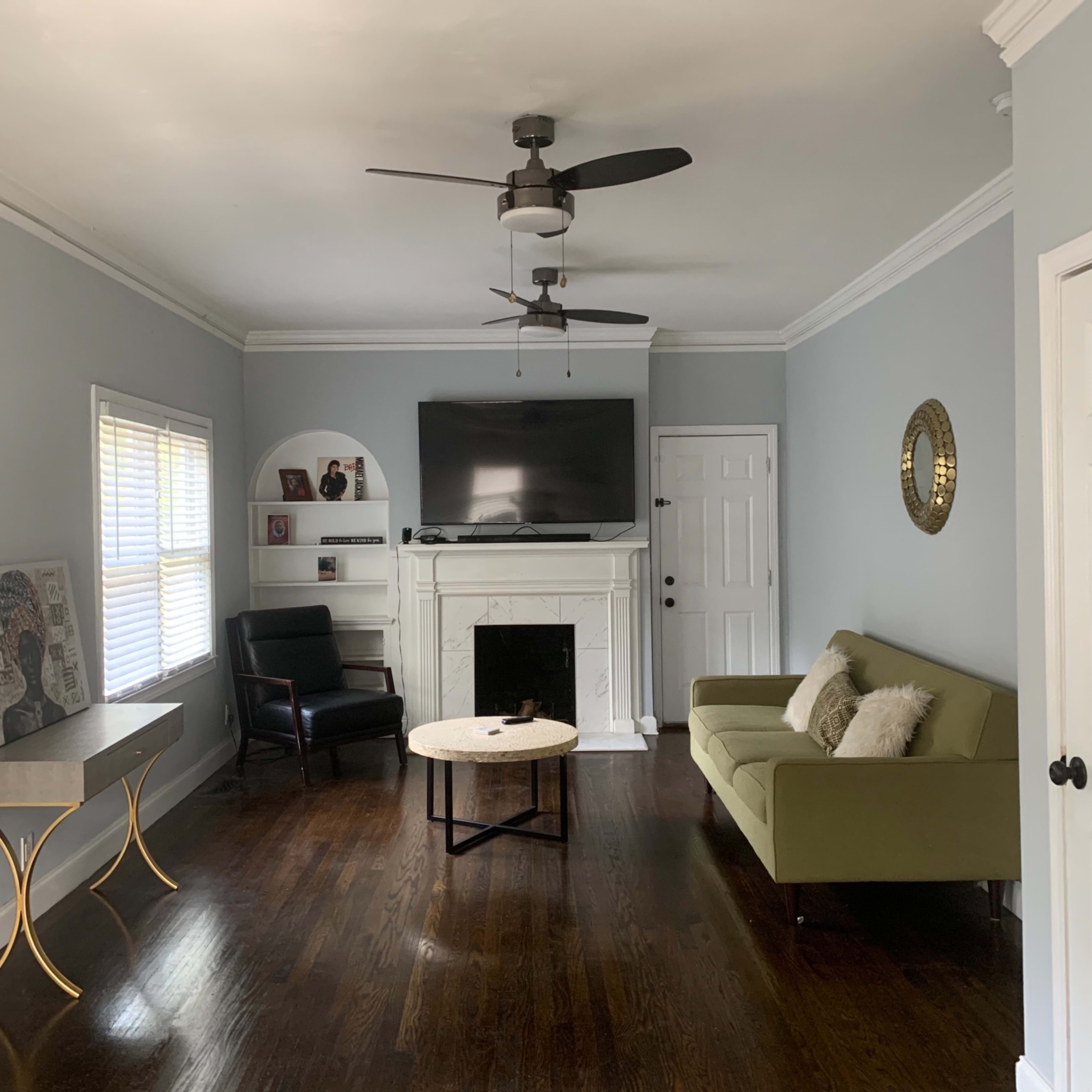 The room features a green sofa, a black chair, a coffee table, and a mounted television above a white fireplace, all set against light gray walls with wooden flooring.