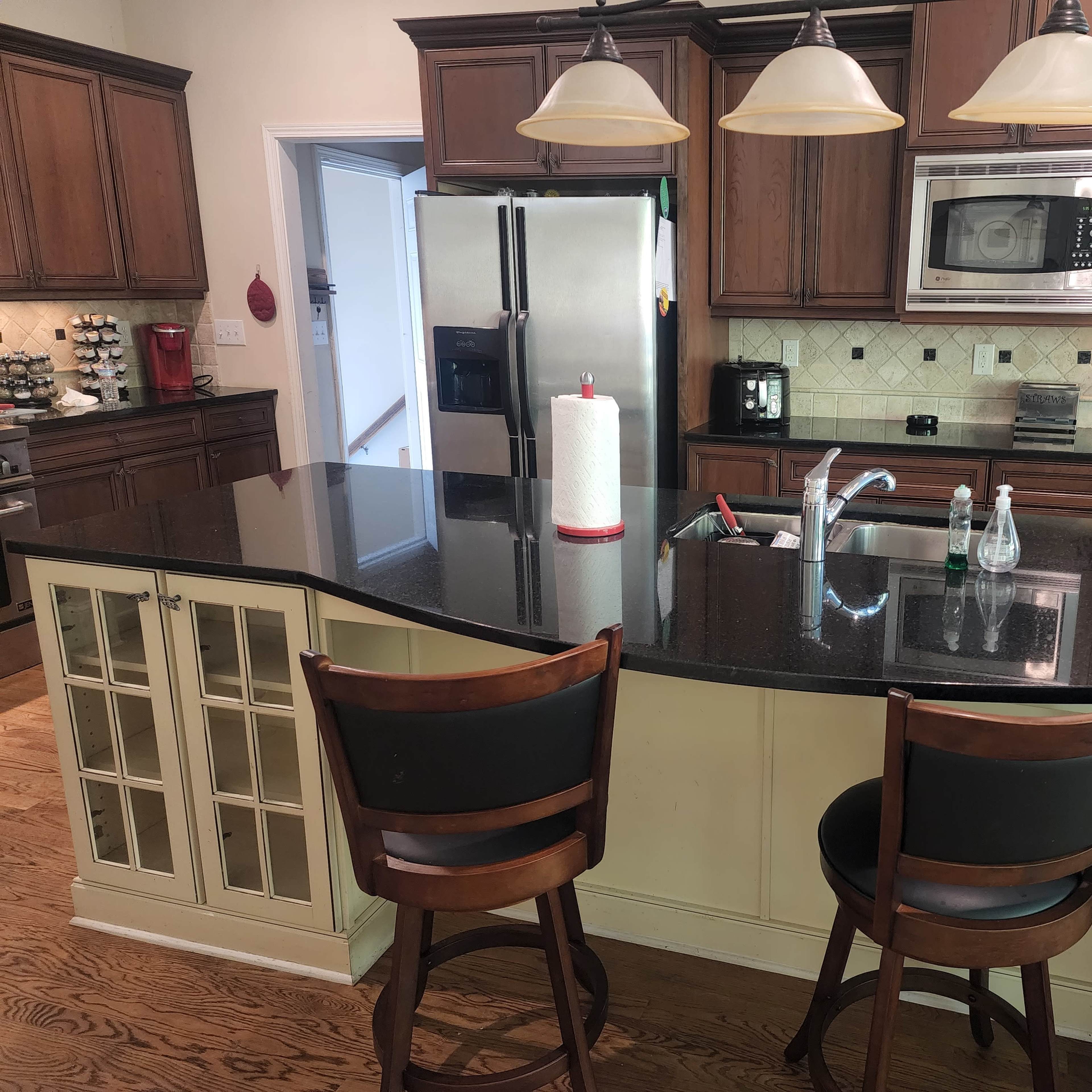 A modern kitchen features dark wooden cabinets, a granite countertop island with two bar stools, and stainless steel appliances.