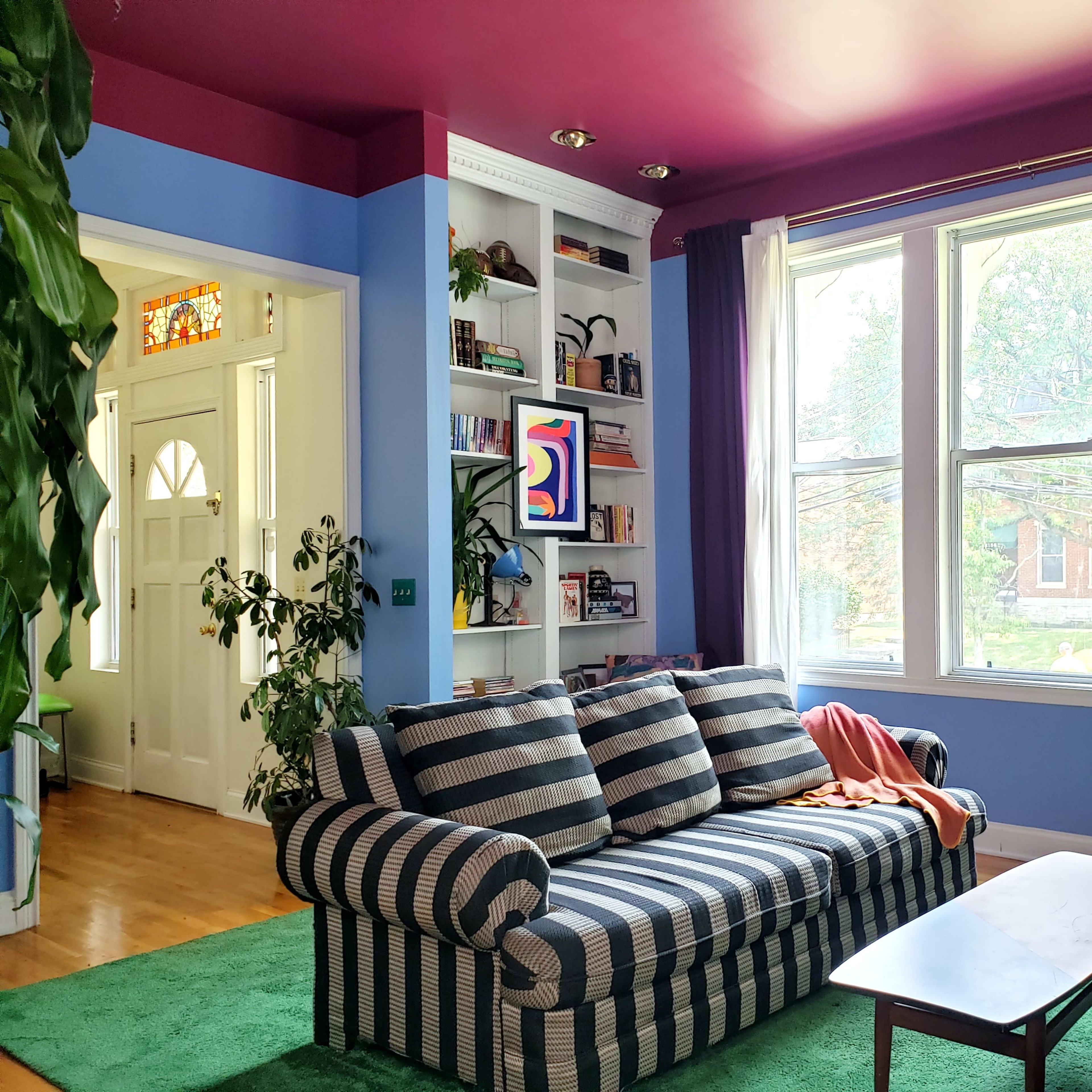 The scene features a cozy living room with a striped couch, a green rug, and large windows surrounded by plants, complemented by colorful walls and shelves.