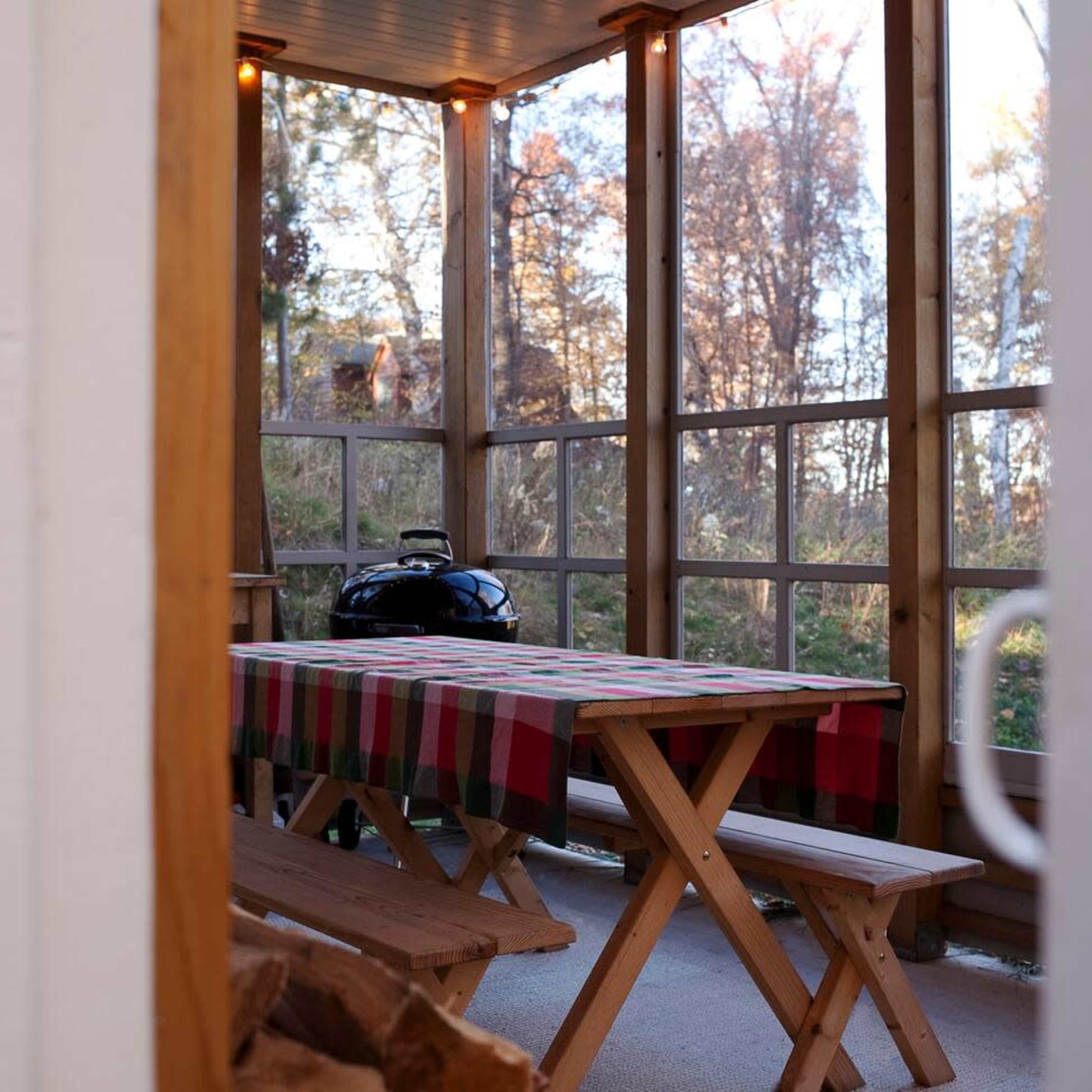 A screened porch features a wooden table covered with a checkered tablecloth and a grill in the background.