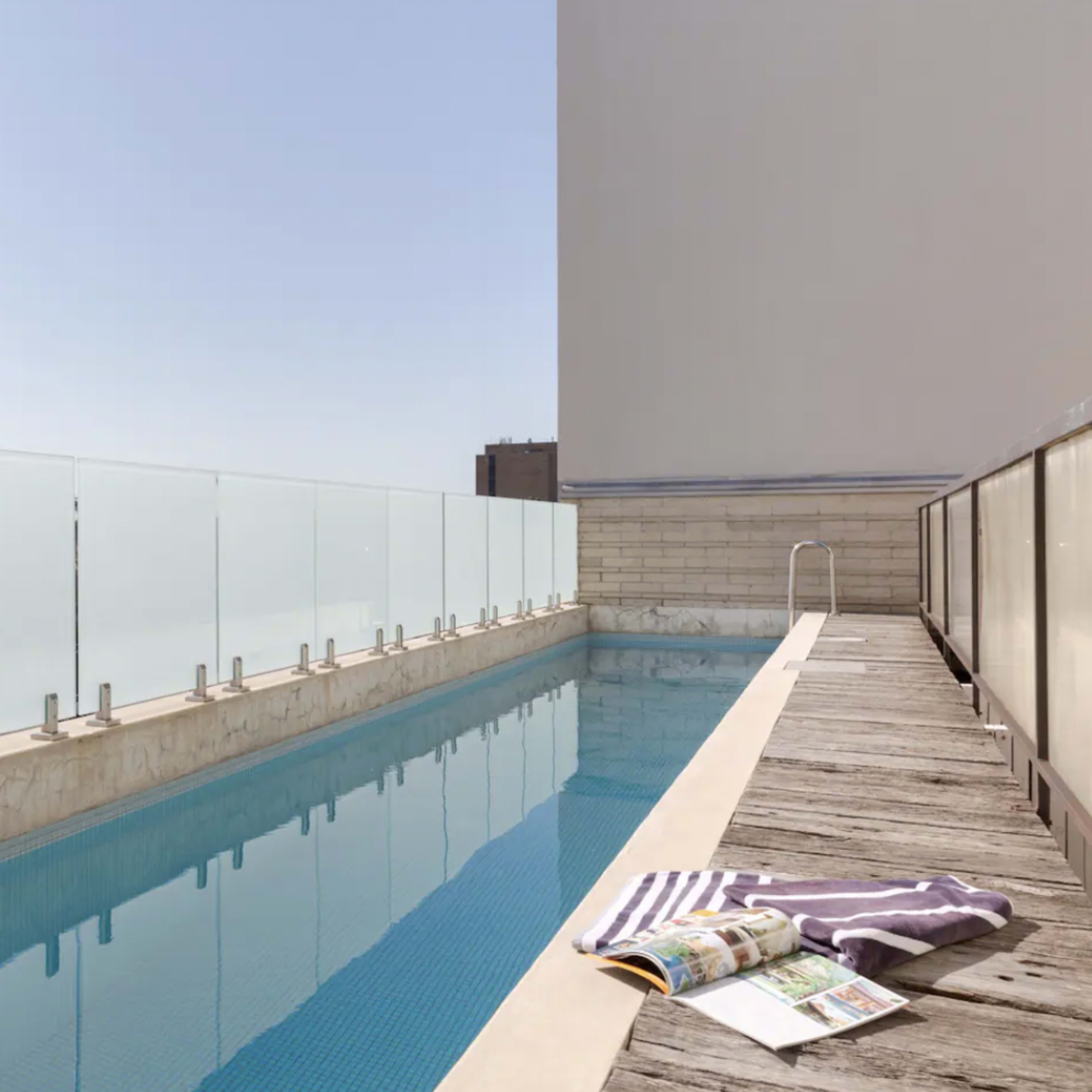 A clear blue swimming pool with a glass railing sits beside a wooden deck under a cloudless sky.