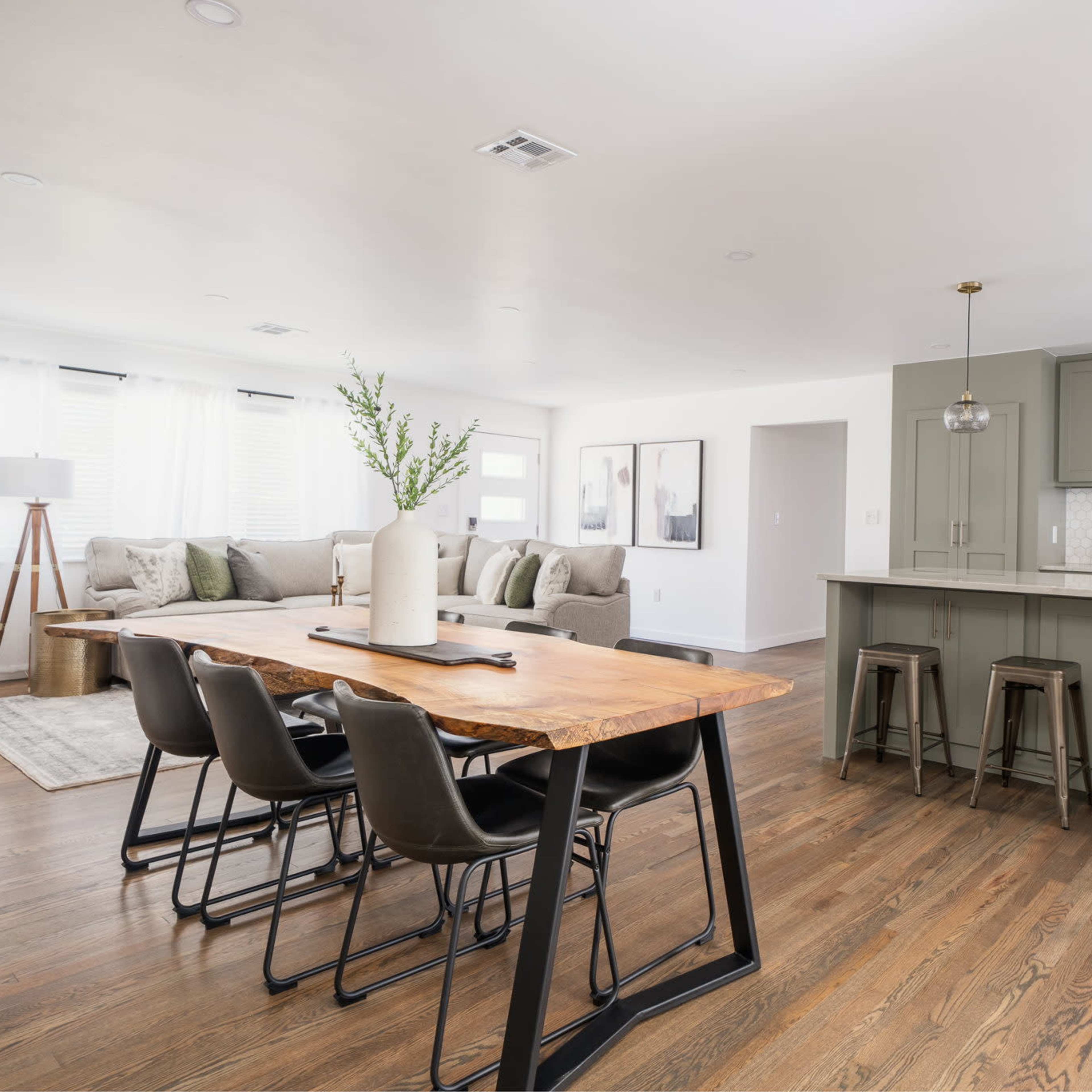 A modern open-concept living space featuring a dining area with a wooden table and black chairs adjacent to a kitchen with green cabinets and a white backsplash.
