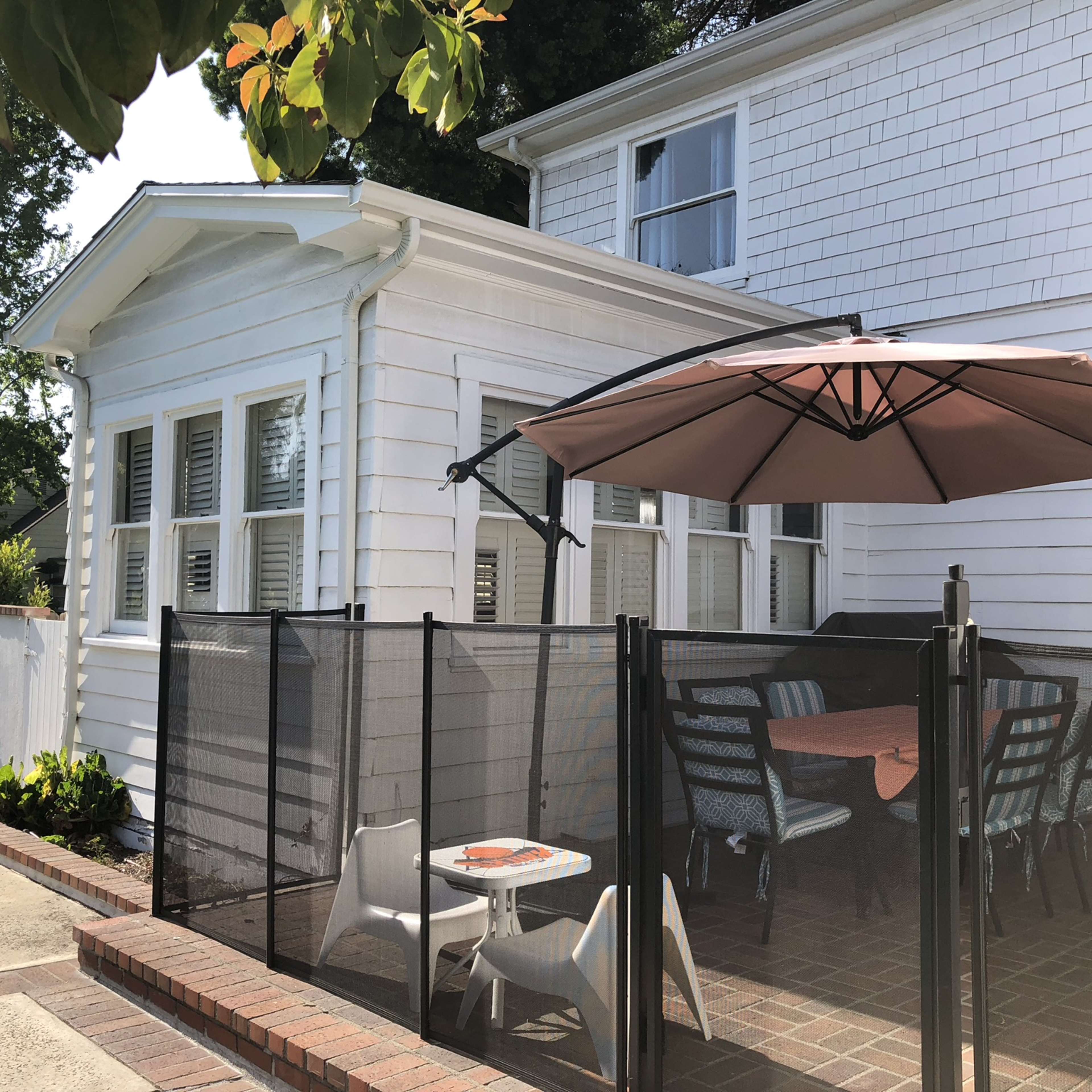 A patio area with a table and chairs, shaded by an umbrella, is located next to a white house with a sunroom.