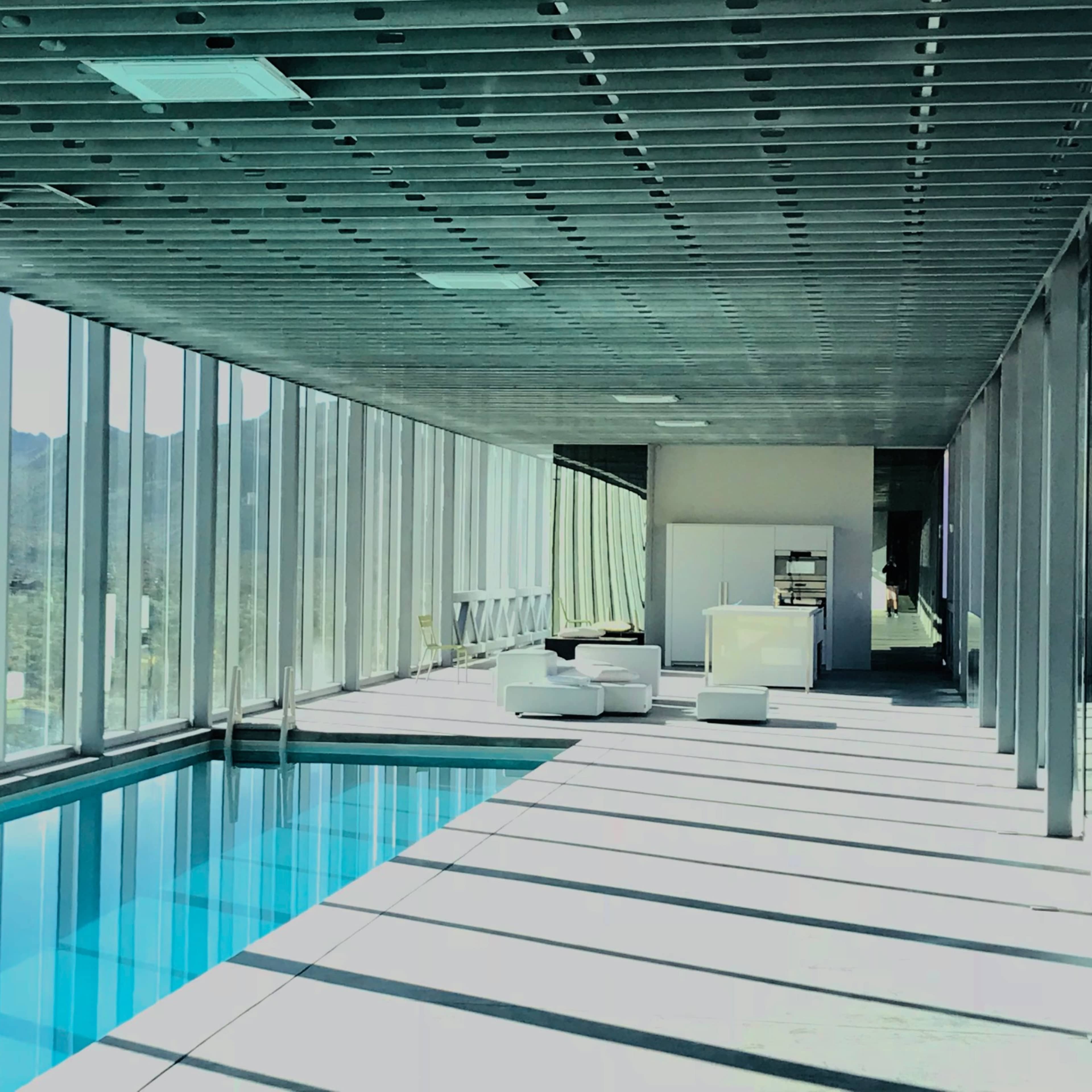 A modern interior space with a floor-to-ceiling glass wall, a swimming pool on one side, and minimalist furniture.