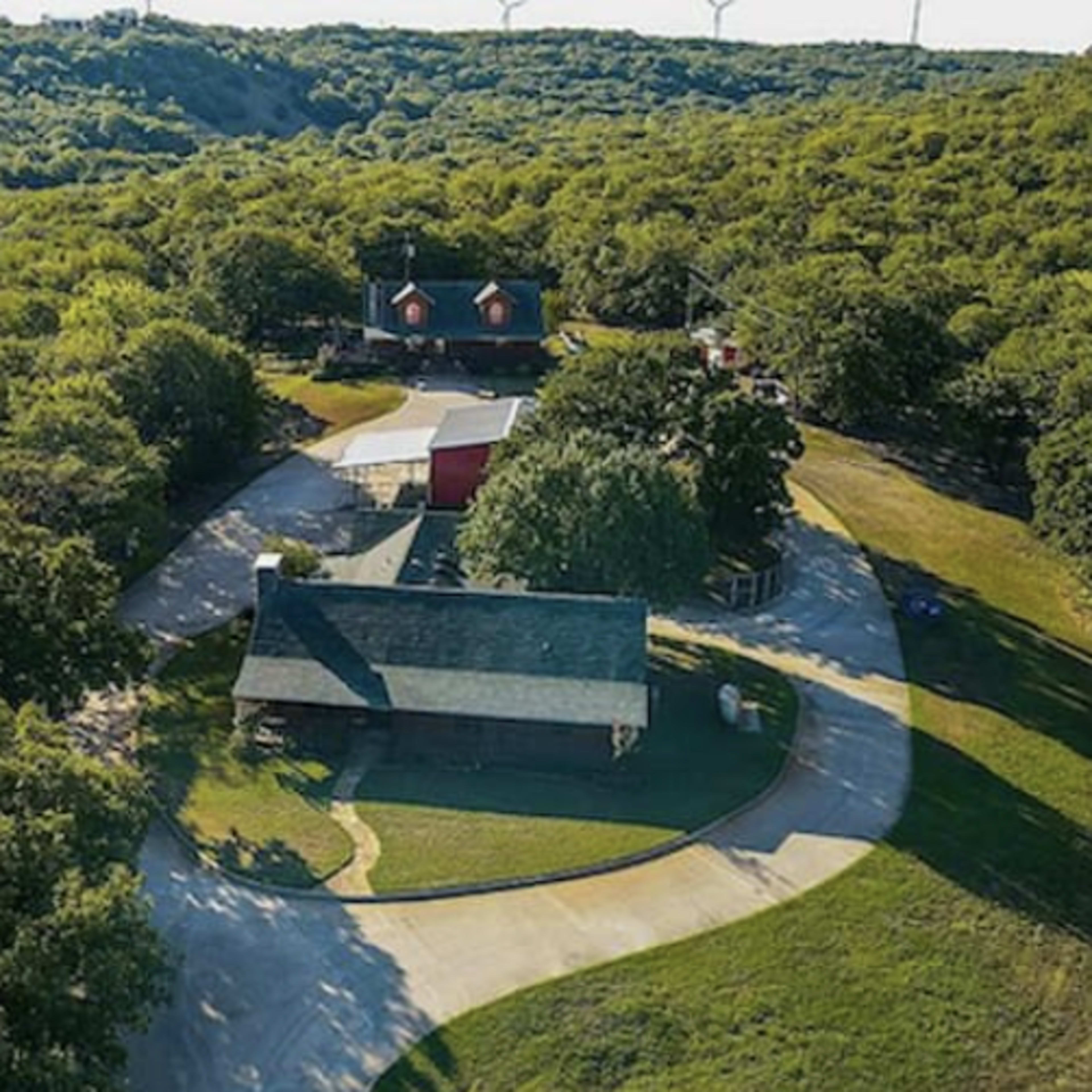 Aerial view of a residential property surrounded by dense green trees, featuring multiple buildings and a winding driveway.