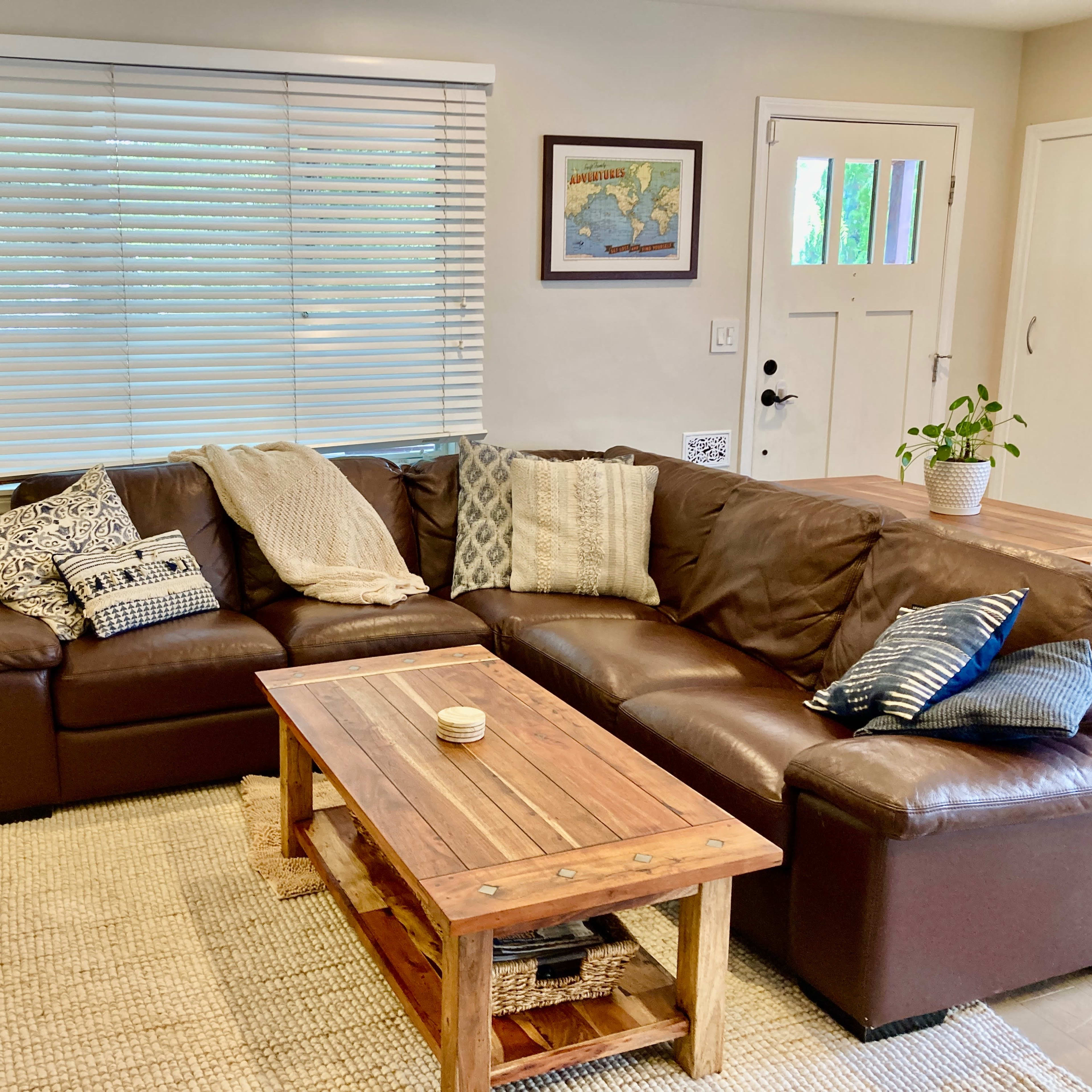 A cozy living room with a sectional brown leather couch, a wooden coffee table, and a small potted plant beside a window covered with blinds.