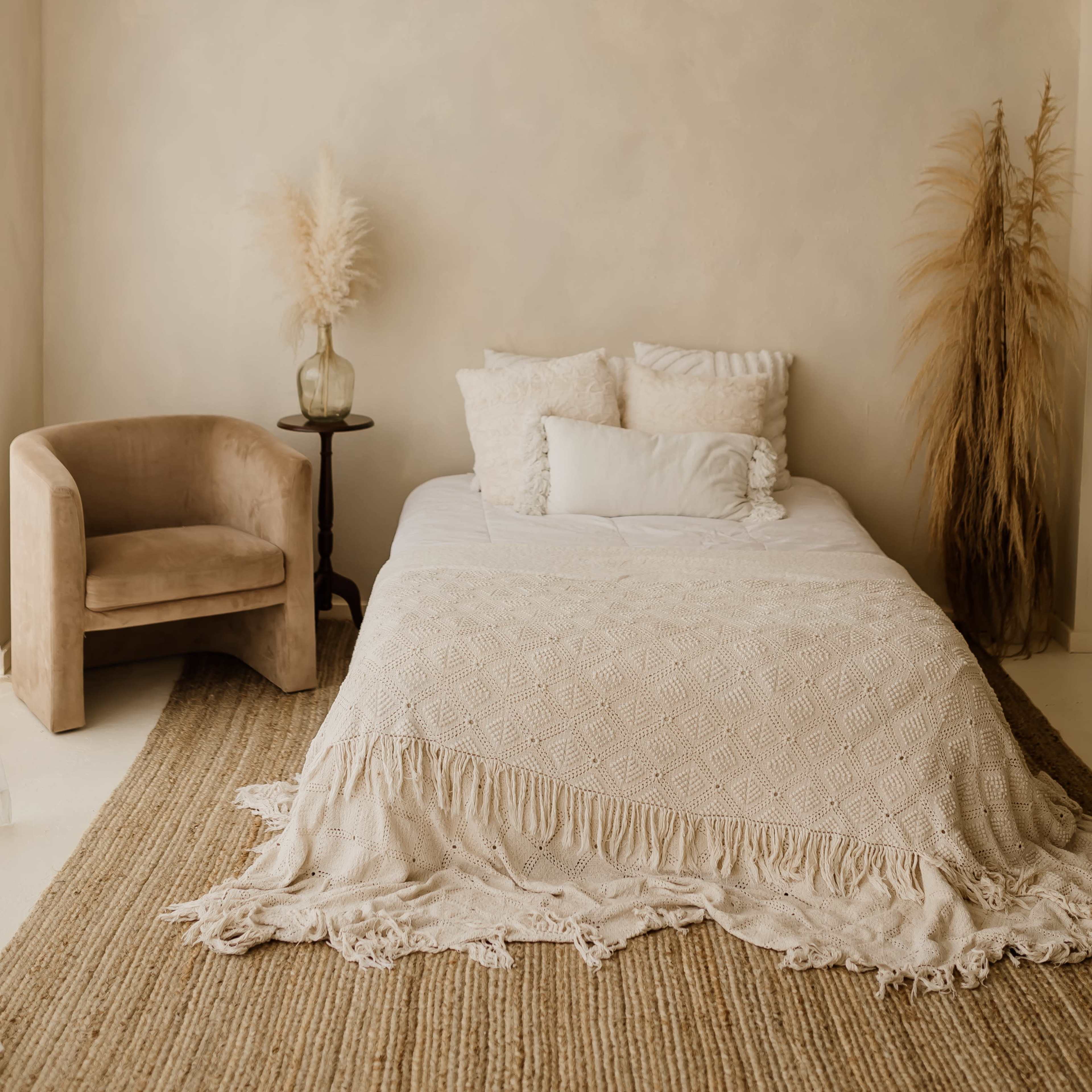 A minimalistic bedroom featuring a bed with a textured blanket, a chair, and decorative dry plants against a neutral wall.