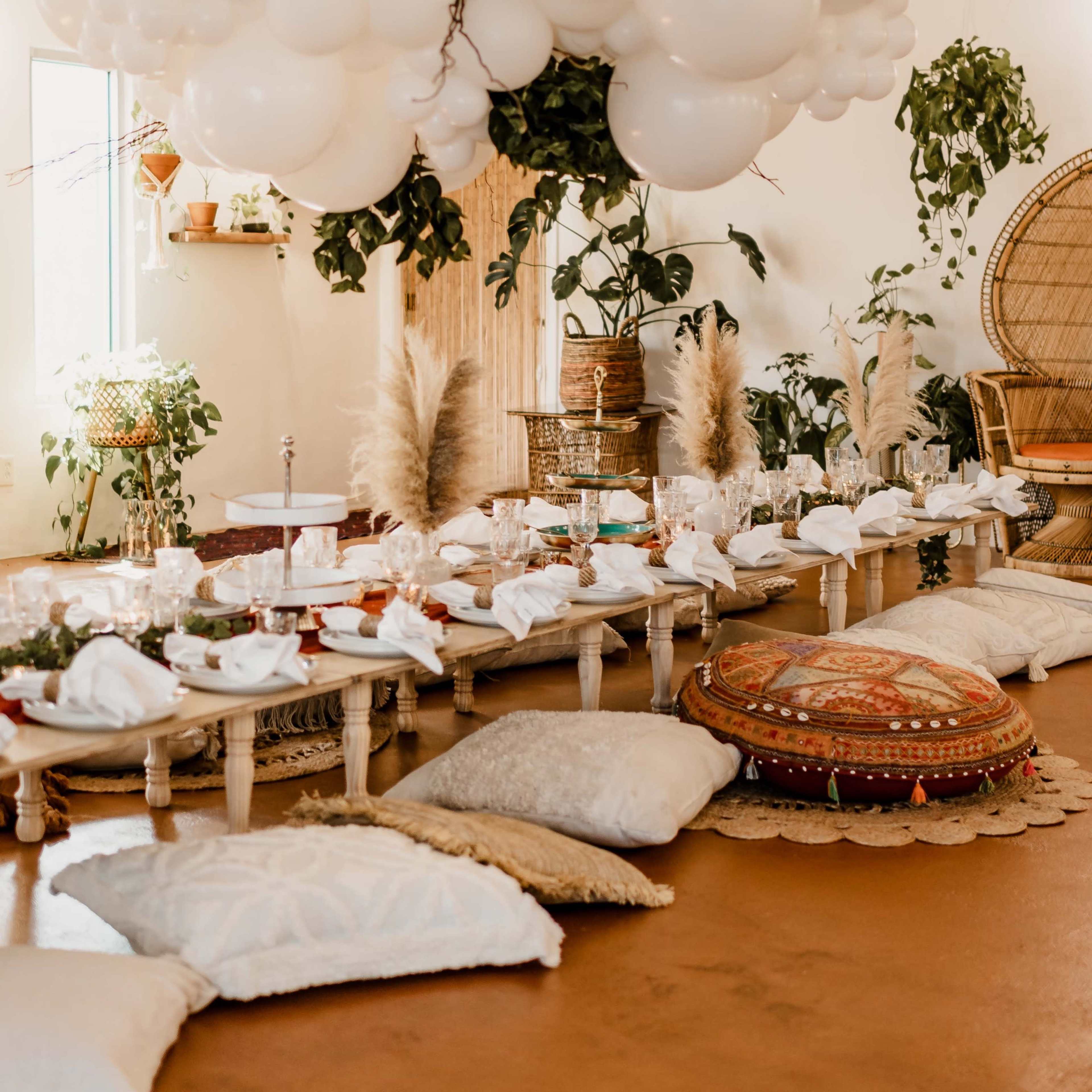 A spacious room features a long table set with plates and napkins, surrounded by decorative cushions and topped with a cloud-like arrangement of white balloons and greenery.