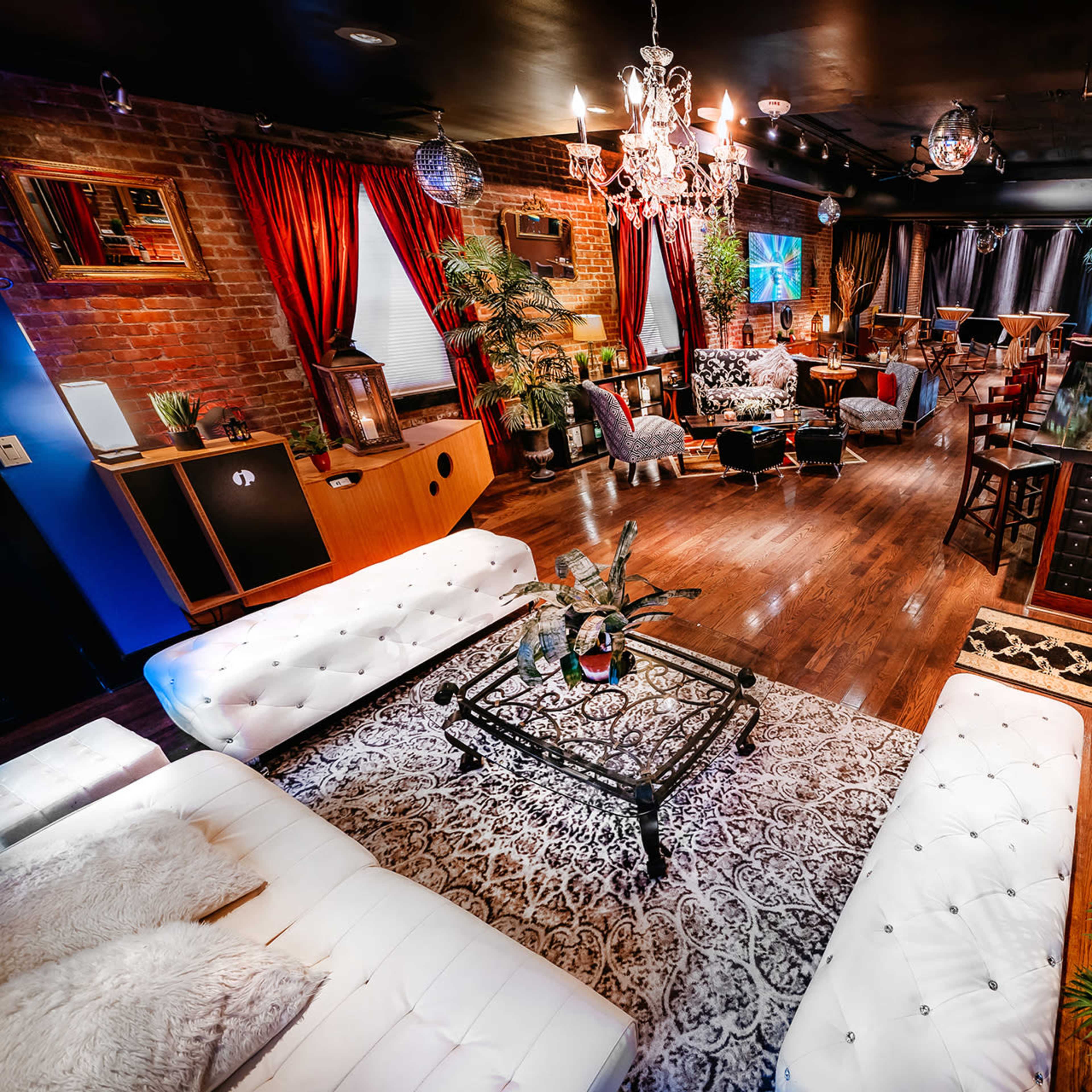 A stylish lounge area filled with plush seating, decorative plants, and a bar, illuminated by chandeliers and accent lighting.