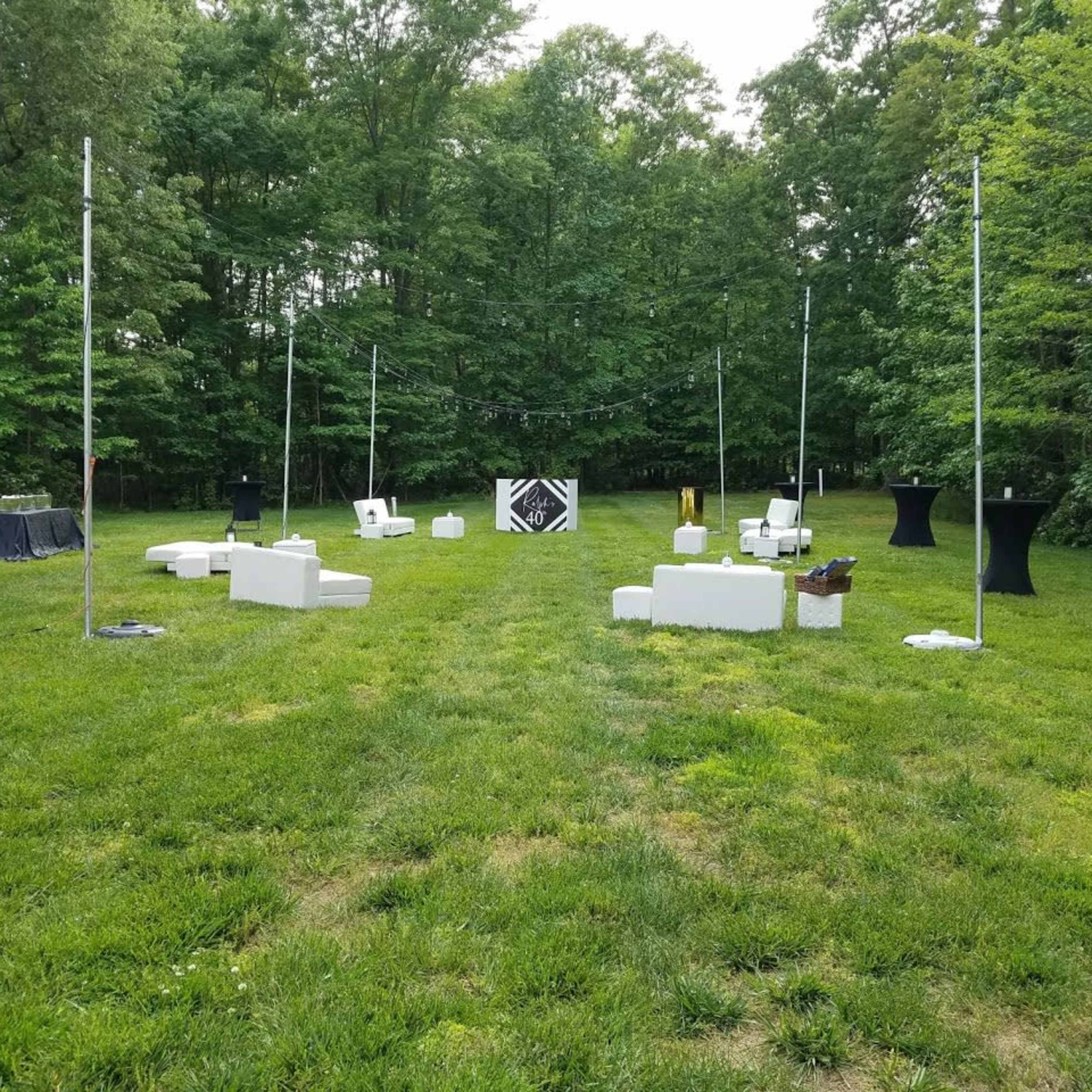 A grassy outdoor area arranged with white seating, black tables, and a central display setup surrounded by tall green trees.
