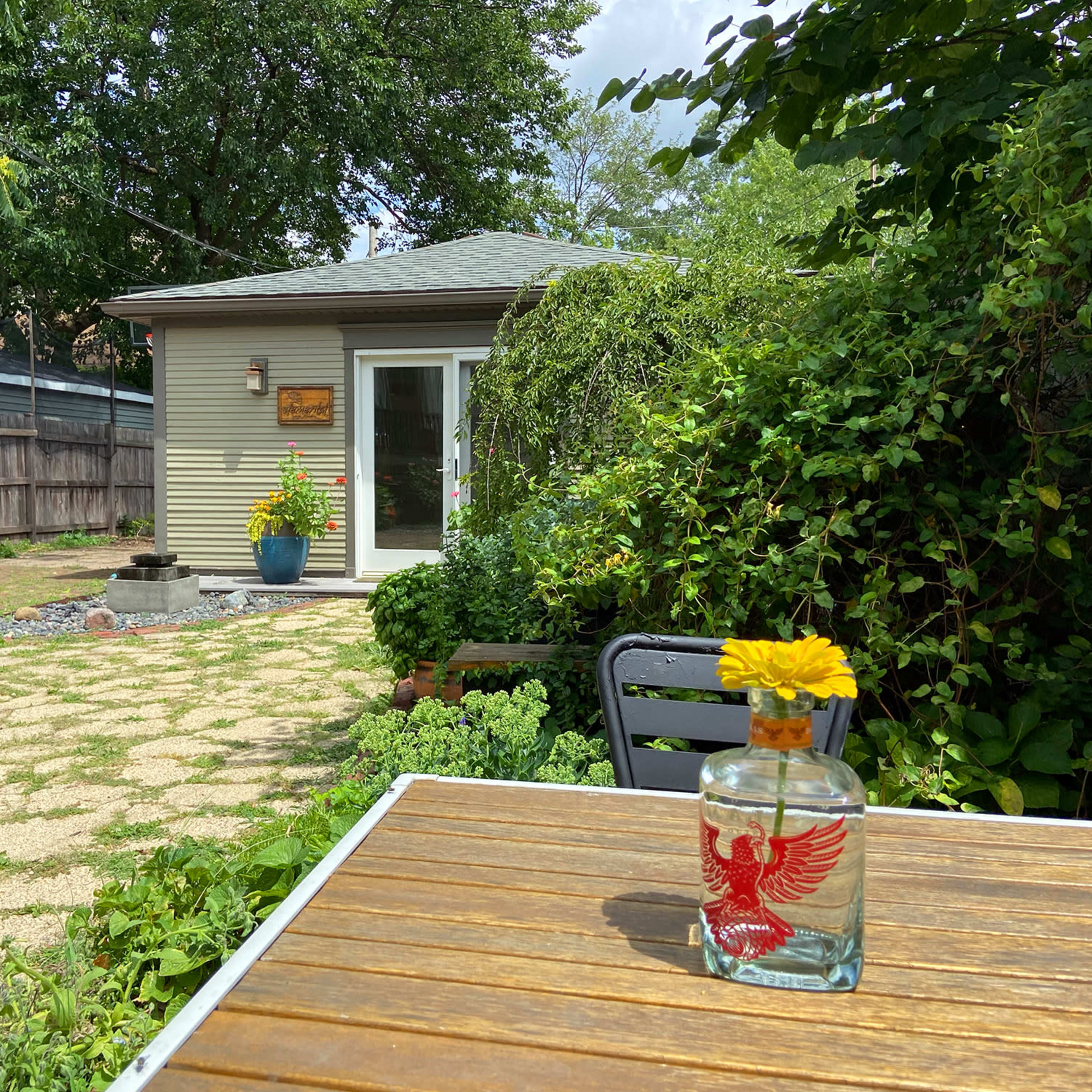 A small garden space features a wooden table with a decorative bottle and a flower, while a house and lush greenery are in the background.