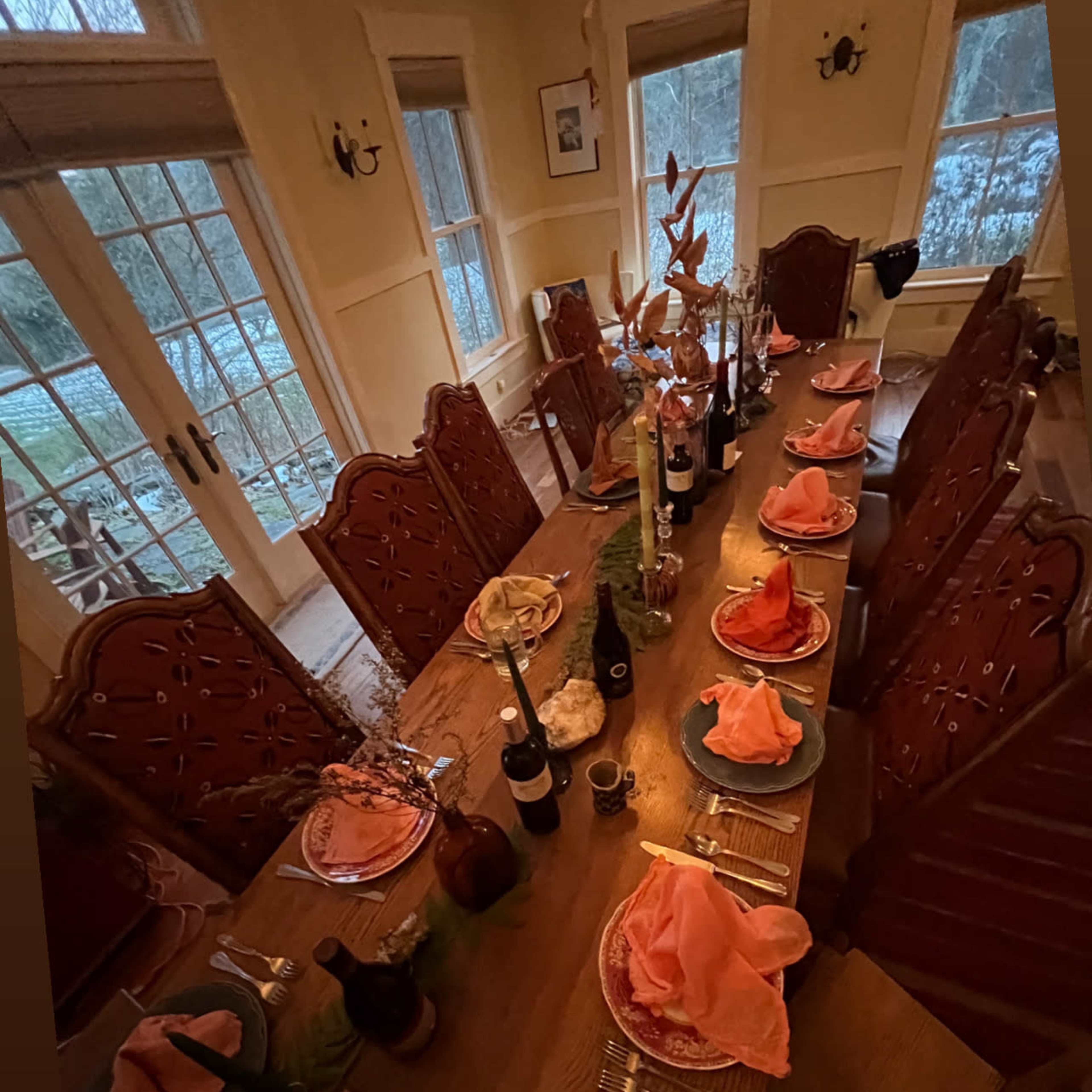 A long wooden dining table is elegantly set with plates, pink napkins, and candles, surrounded by intricately designed chairs in a well-lit room with large windows overlooking a garden.
