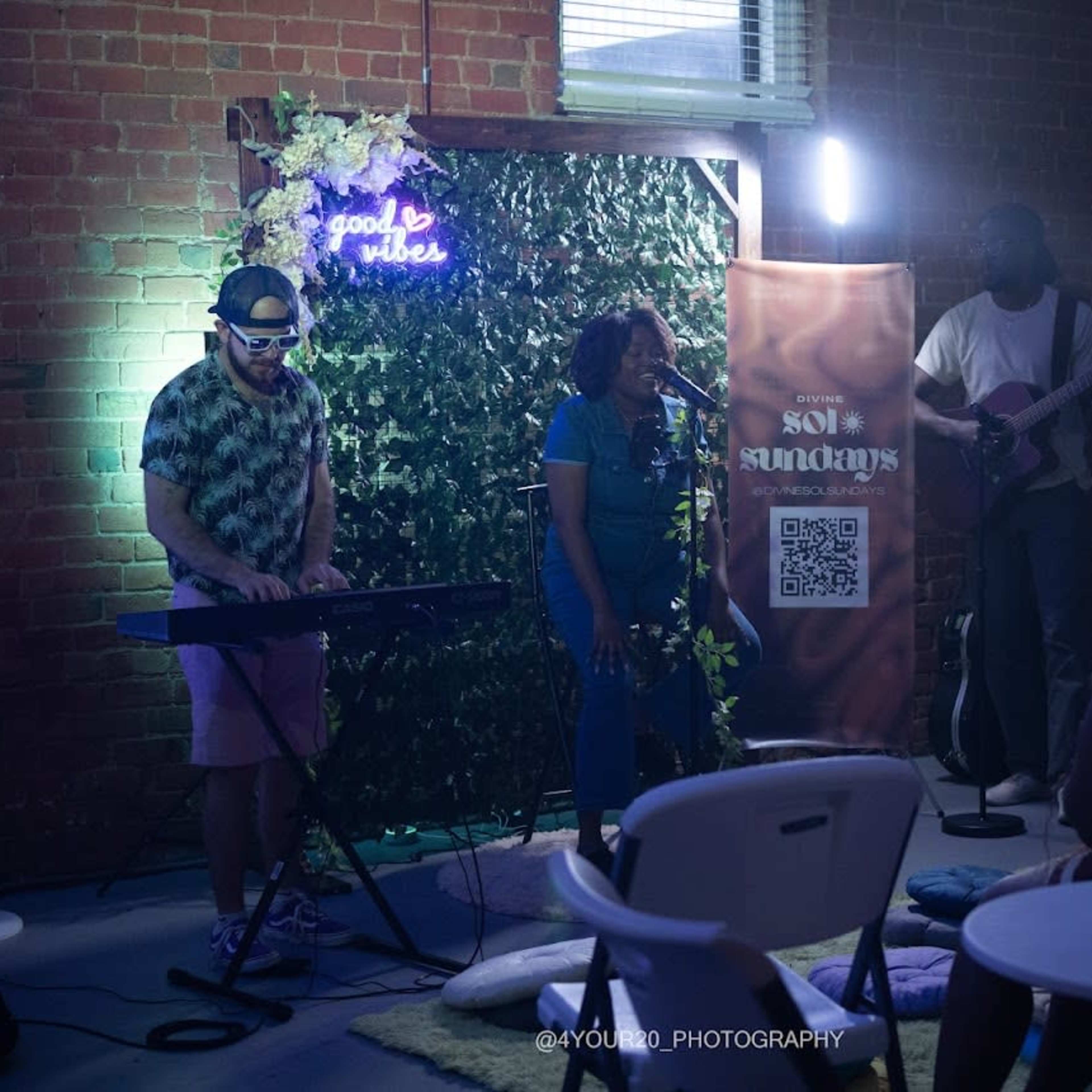 A singer performs on stage while accompanied by a keyboardist and a guitarist in a dimly lit venue decorated with greenery.