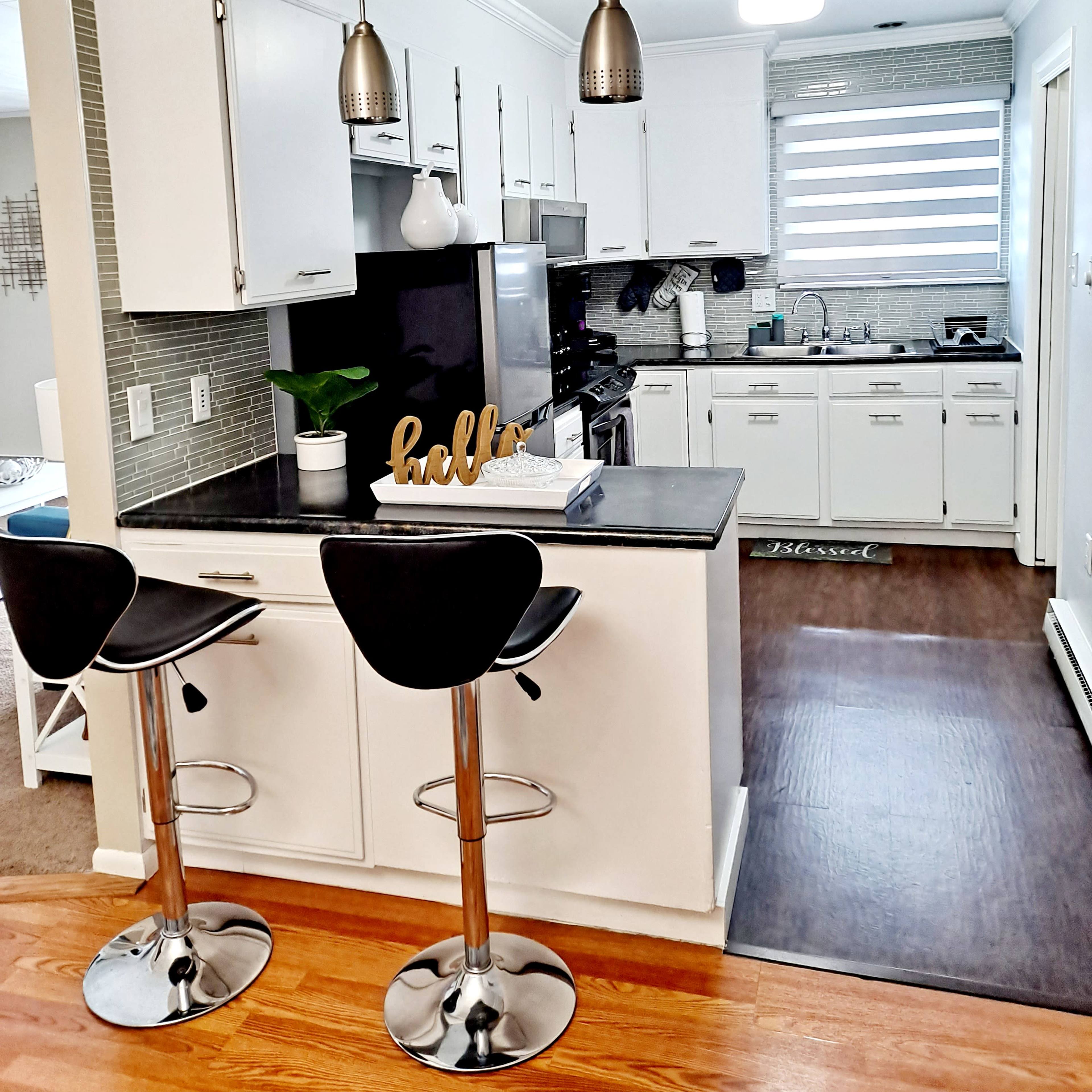 A modern kitchen features white cabinetry, stainless steel appliances, and two bar stools at a black countertop, with pendant lighting above.