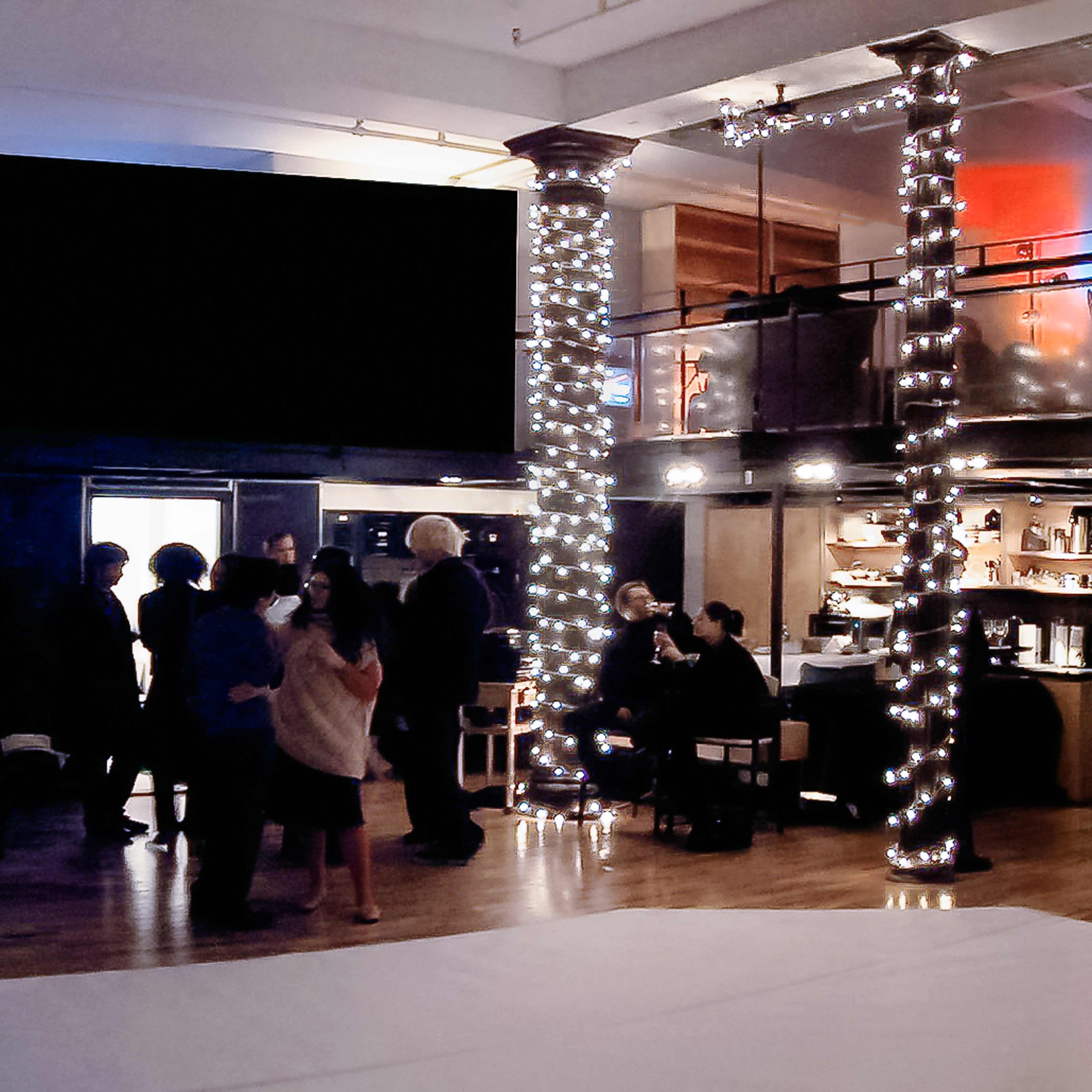 A spacious interior with multiple levels, decorated with string lights, where people are gathered for an event.