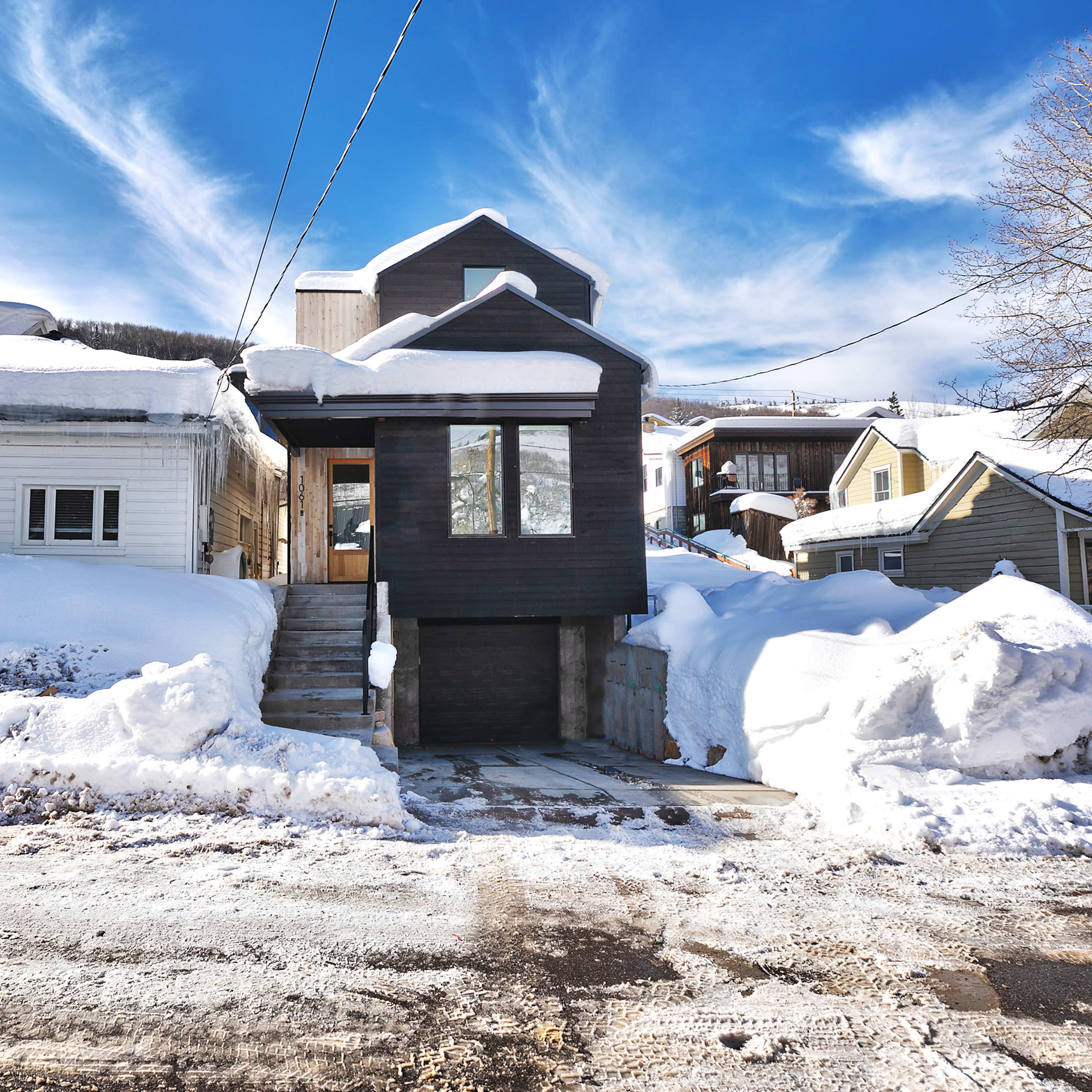 A modern two-story house with a dark exterior design is situated among snow-covered properties against a blue sky with wispy clouds.