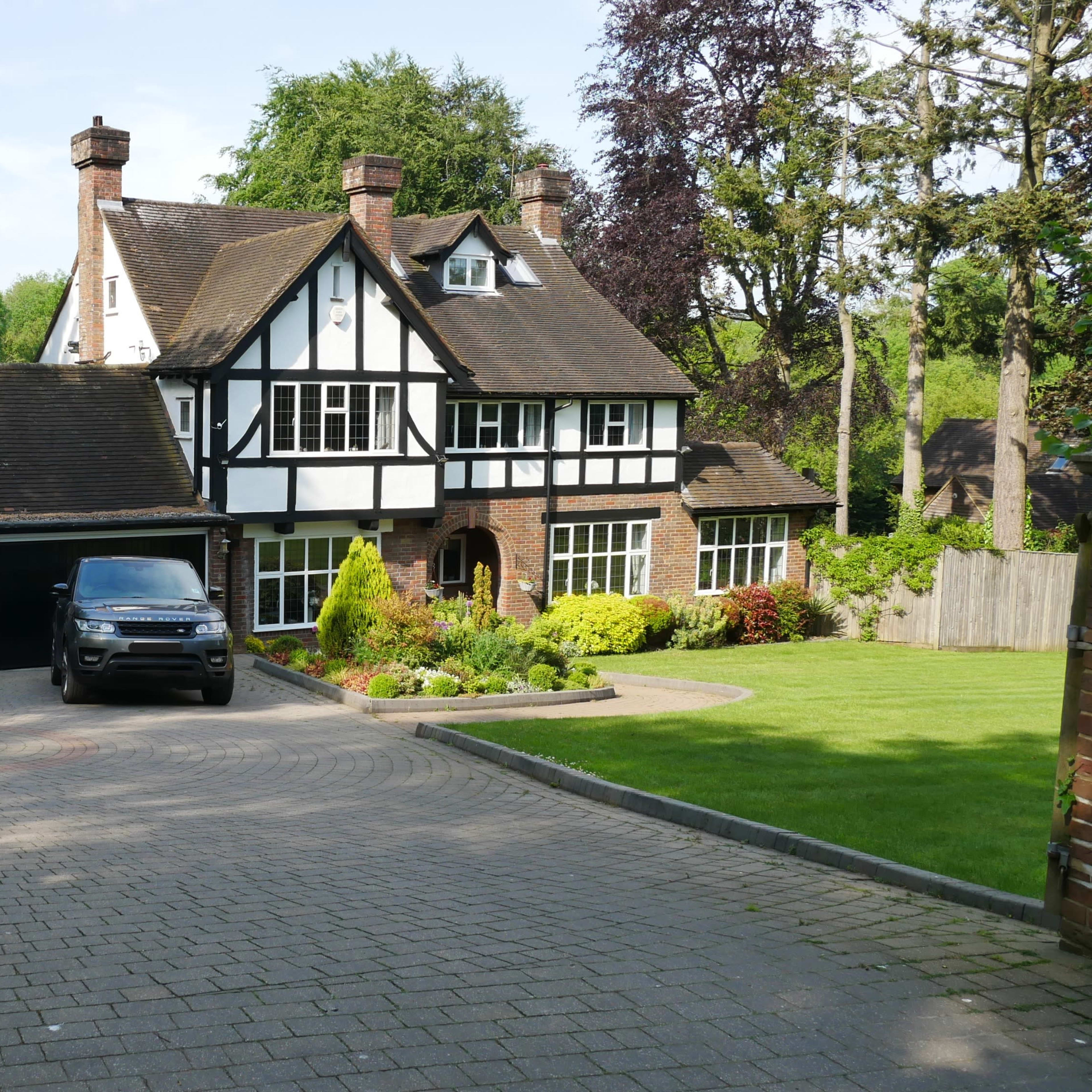 A large, two-story house with a black and white Tudor-style facade, two cars parked in the driveway, and a well-maintained garden surrounding the property.