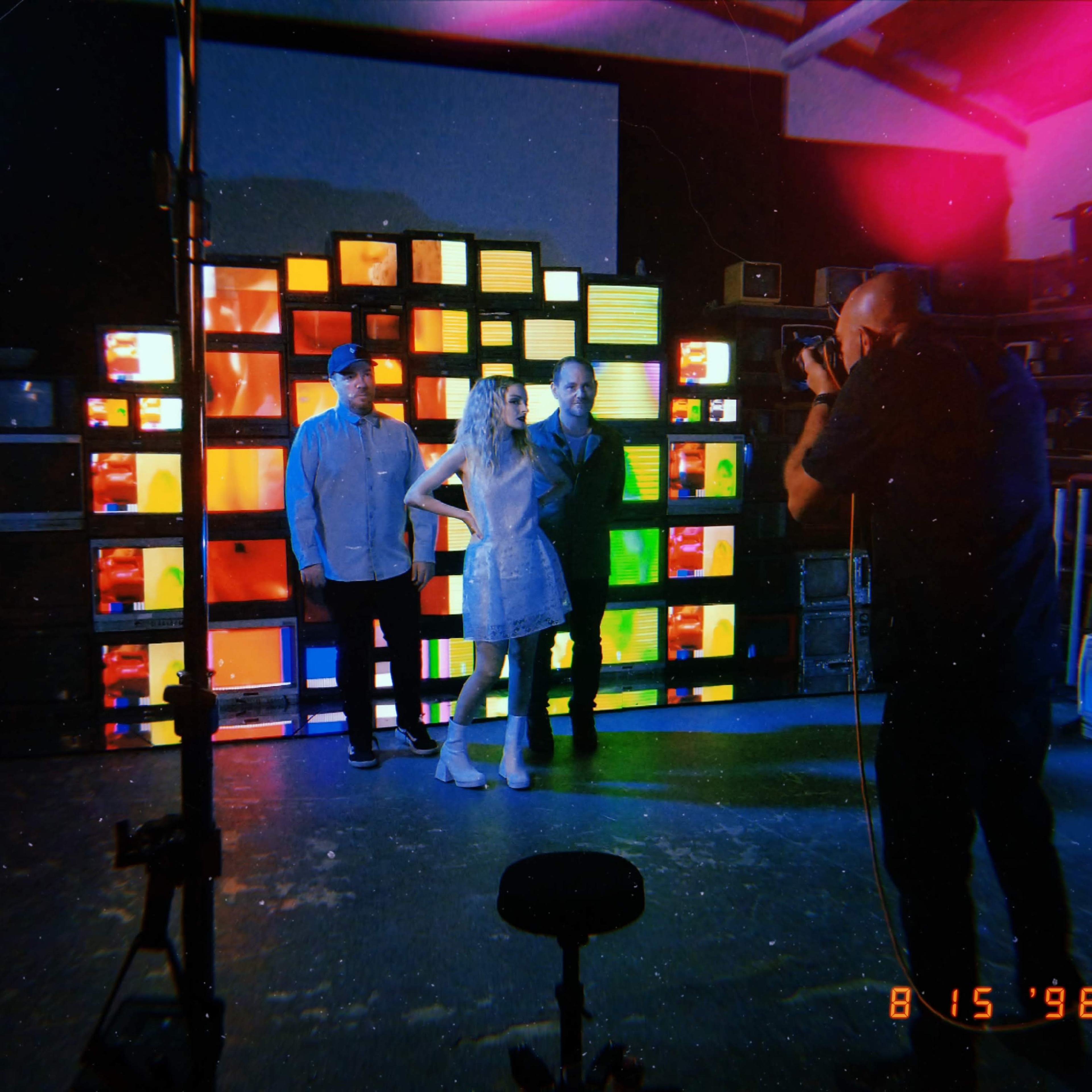 A photographer captures a posed shot of three individuals in front of a backdrop made of colorful television screens in a dimly lit studio.
