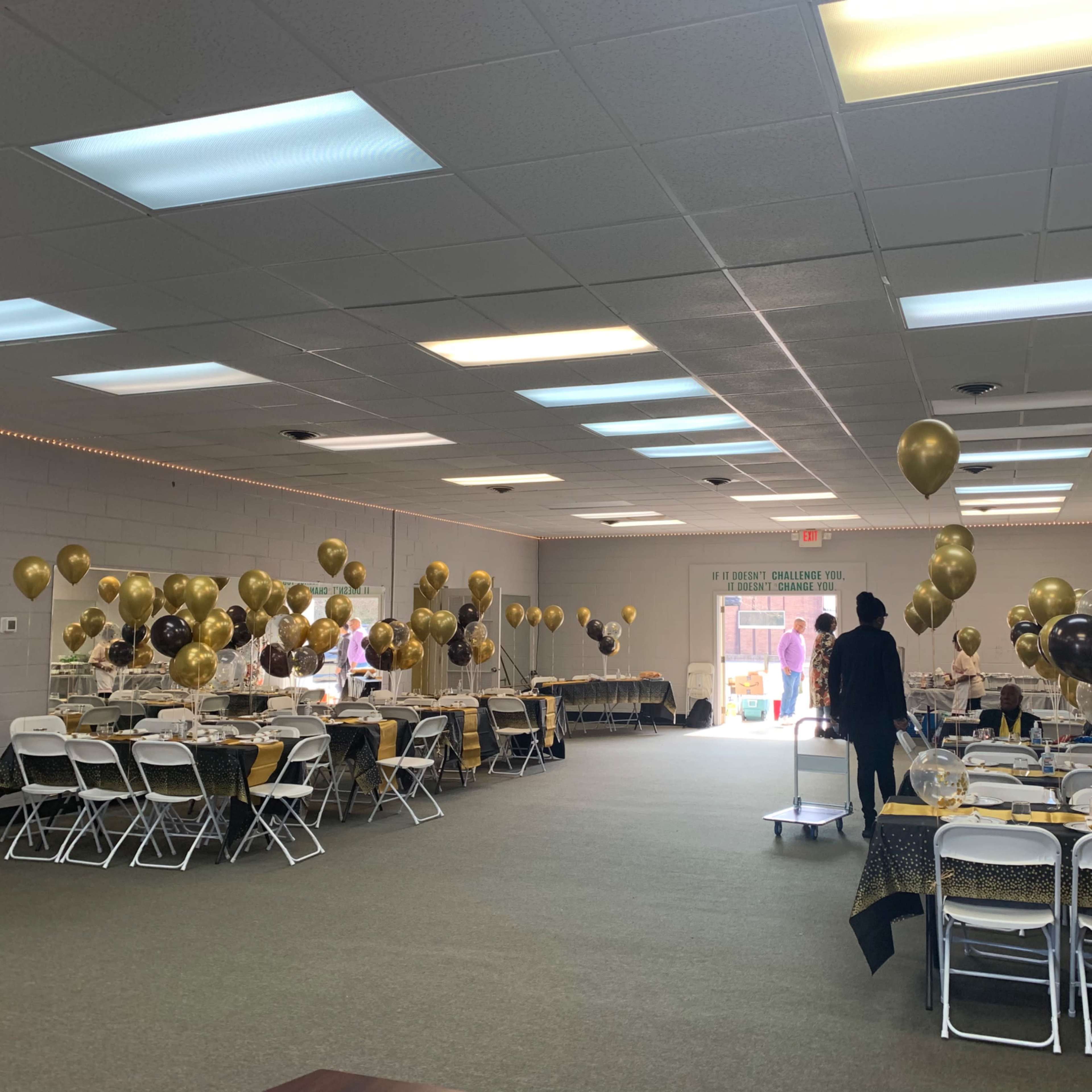 A spacious event room is decorated with gold and black balloons, and tables are arranged for a celebration.
