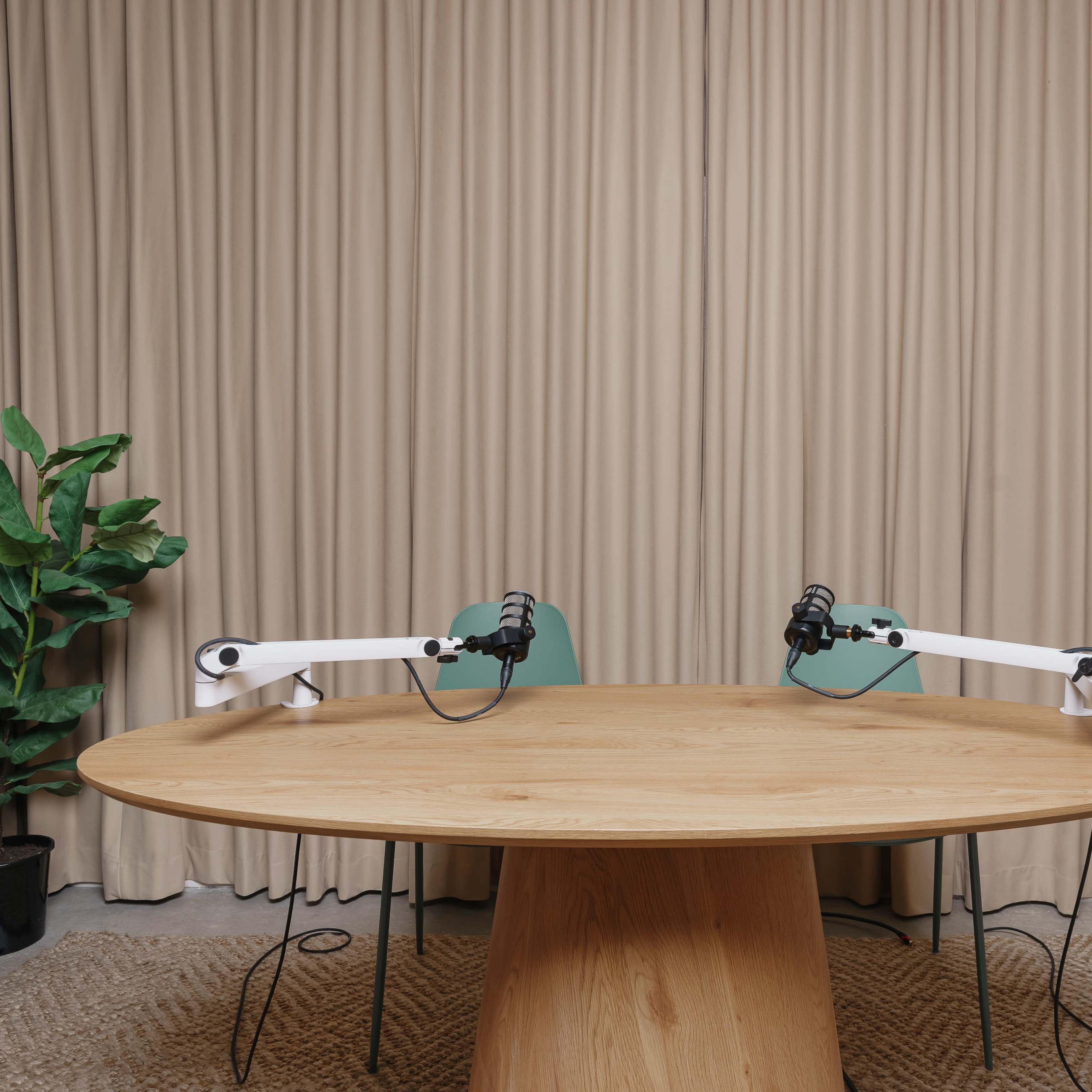 A podcast studio setup with a large round table, two chairs, and microphone stands against a backdrop of beige curtains and potted plants.